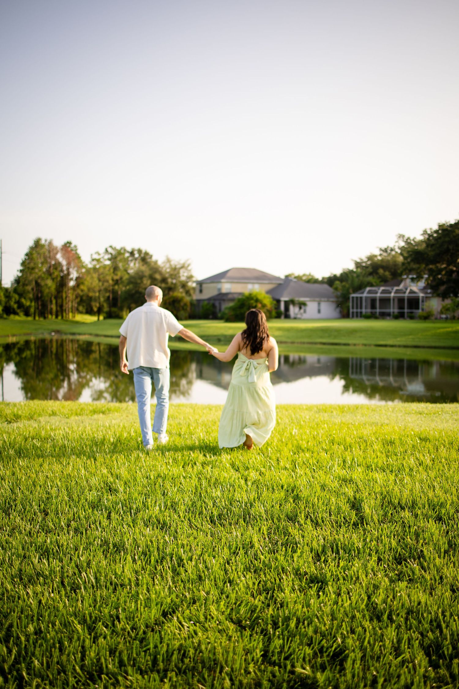 Couple holding hands walking on lush green lawn beside a tranquil pond at sunset with houses in background.