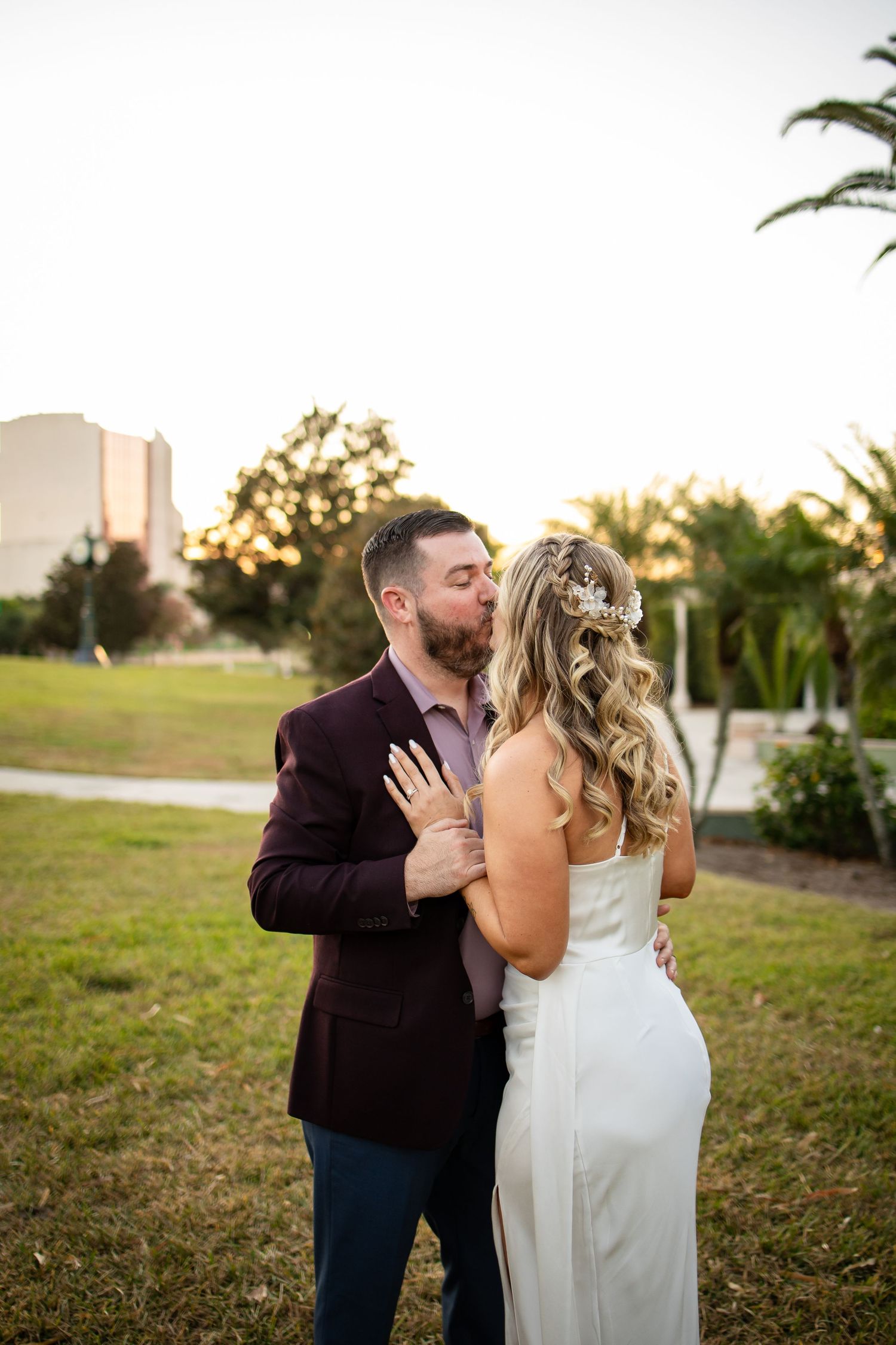 A couple embraces outdoors at sunset with palm trees and a building in the background.