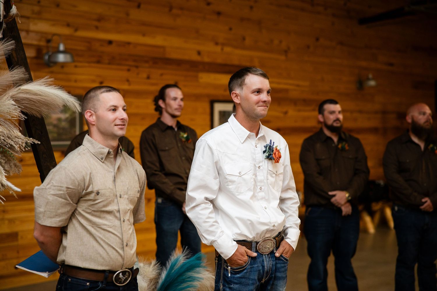 Groomsmen in brown shirts stand smiling inside a rustic wood-paneled wedding venue.