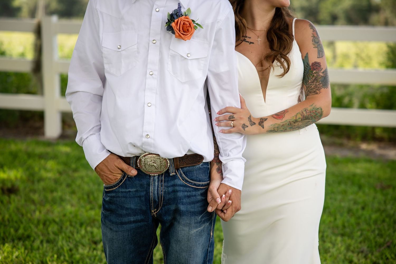 Bride and groom holding hands, groom in white shirt with boutonniere and jeans, bride in white gown with tattoos.
