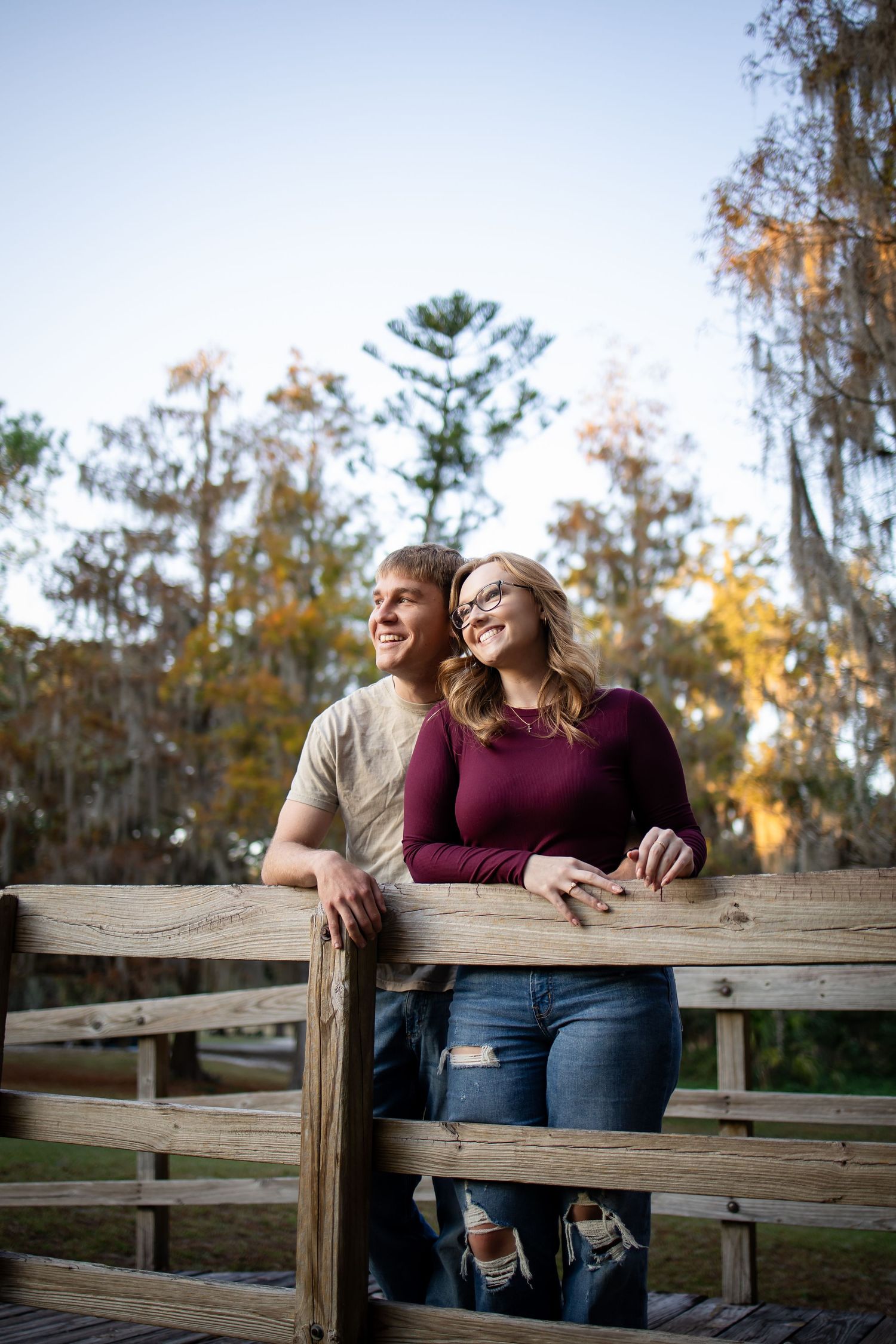 A romantic couple leans against a wooden fence during golden hour in a park setting.