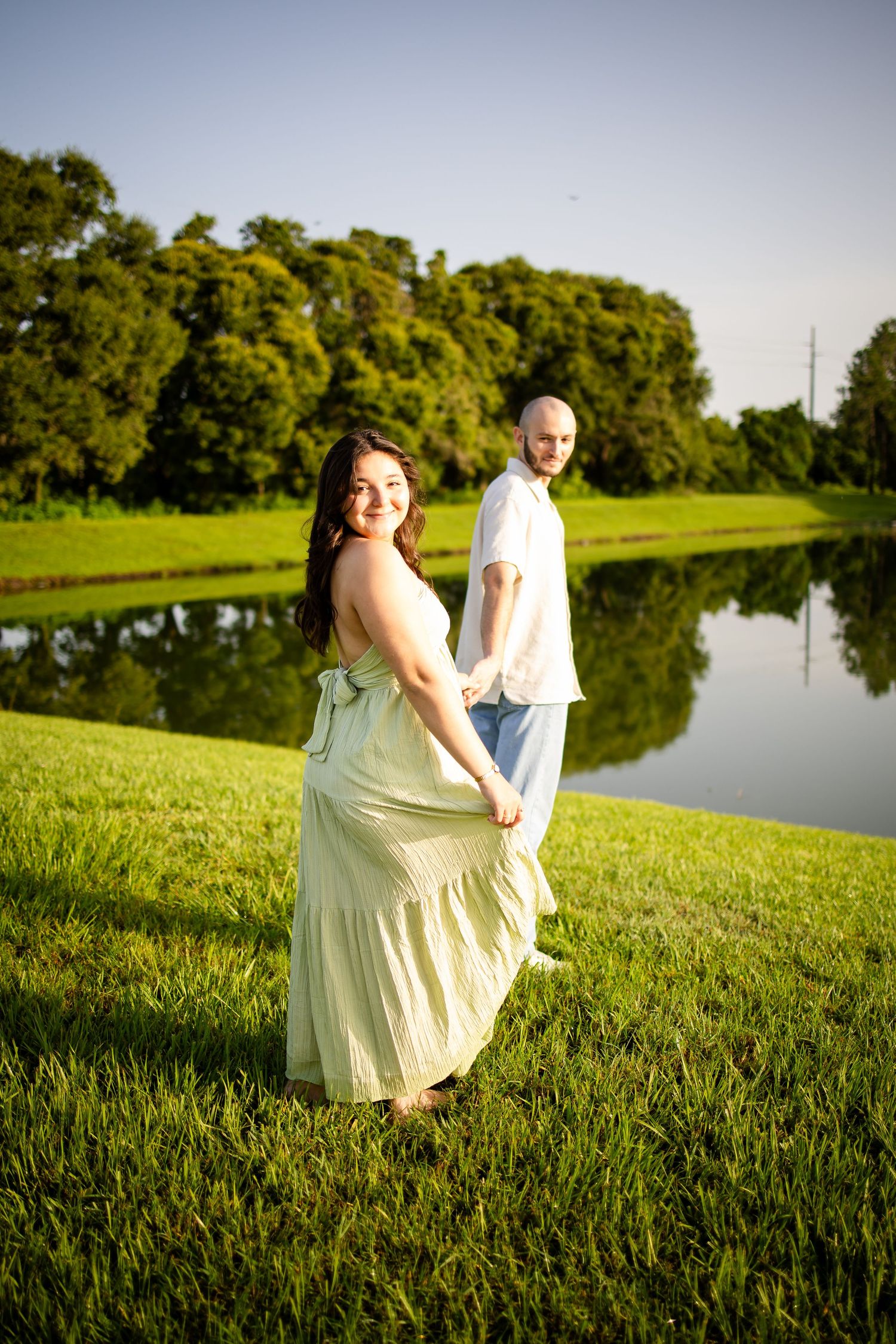 Two people in light flowy dresses sit together on grassy hill by serene lake during golden hour sunset.