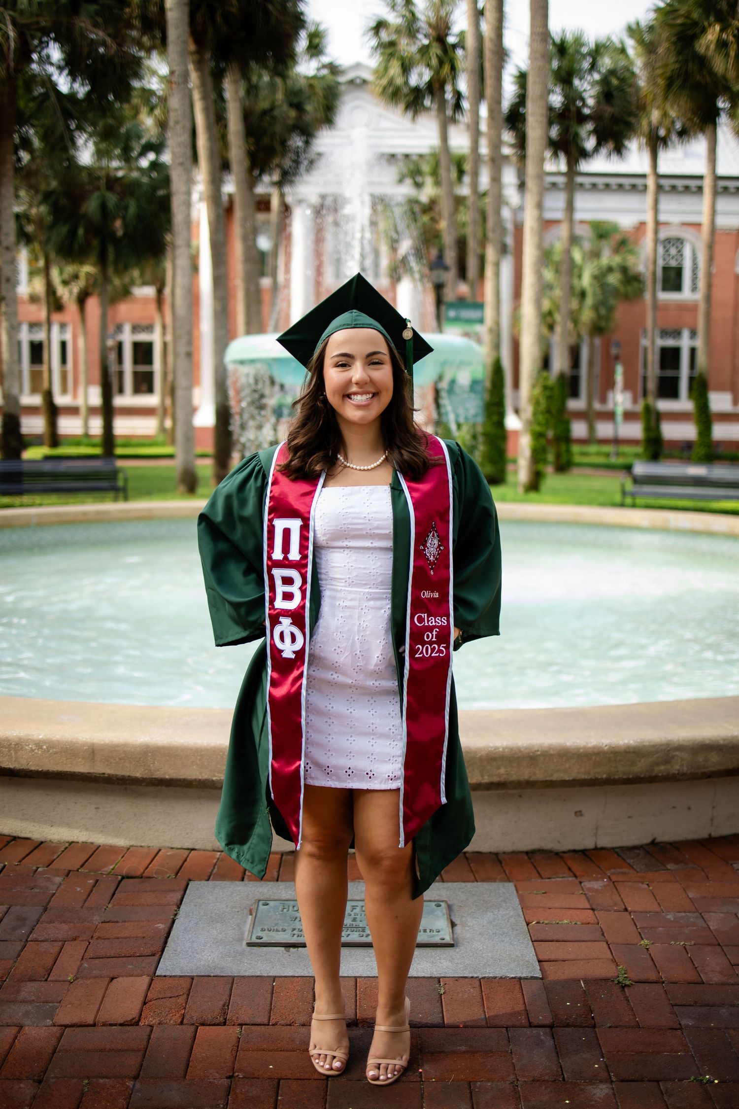 A graduating student wearing a green graduation gown and honor cords poses by a fountain at a university campus.