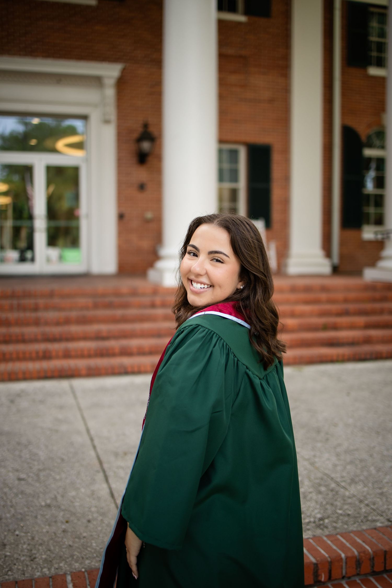 Graduate in green cap and gown smiles over shoulder on brick steps of traditional college building with white columns.