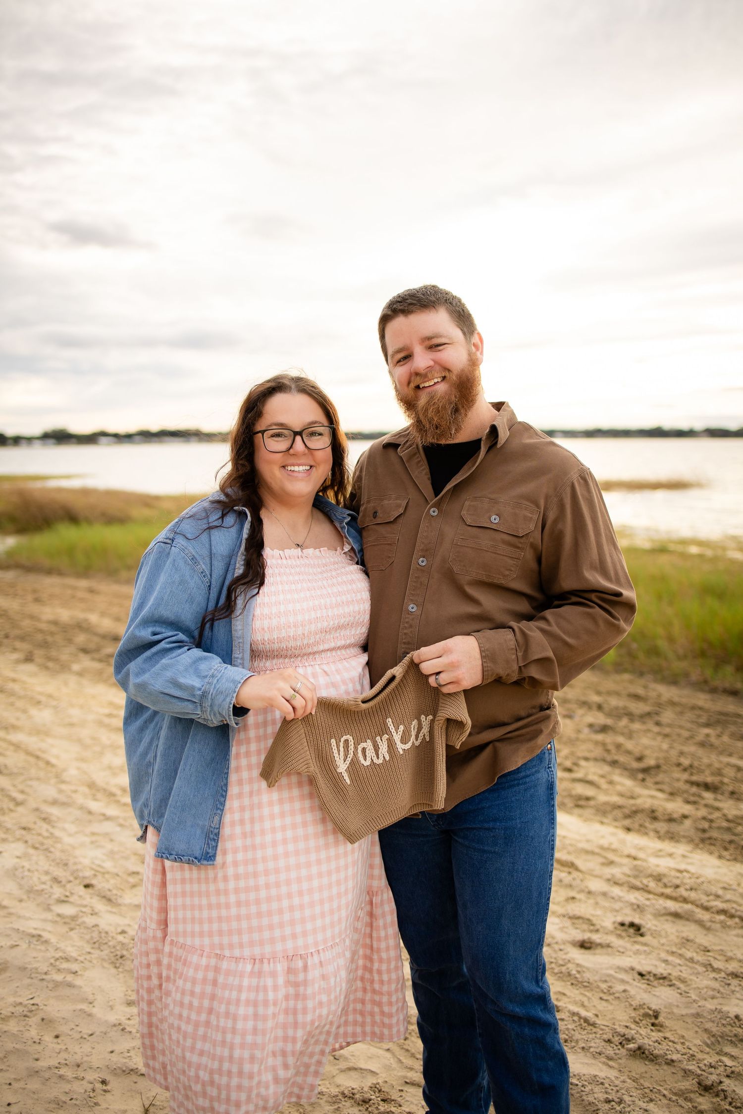 Couple posing together on a beach at sunset with one wearing a pink gingham dress and denim jacket.