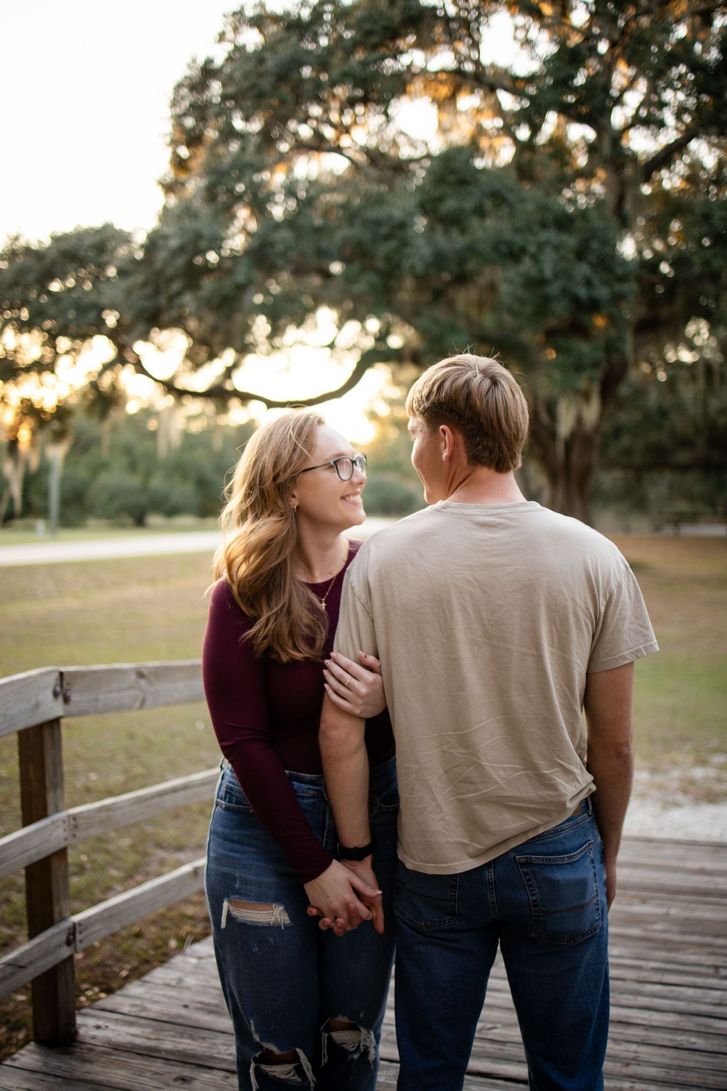 A couple stands on a wooden boardwalk during sunset with Spanish moss draped trees in the background.