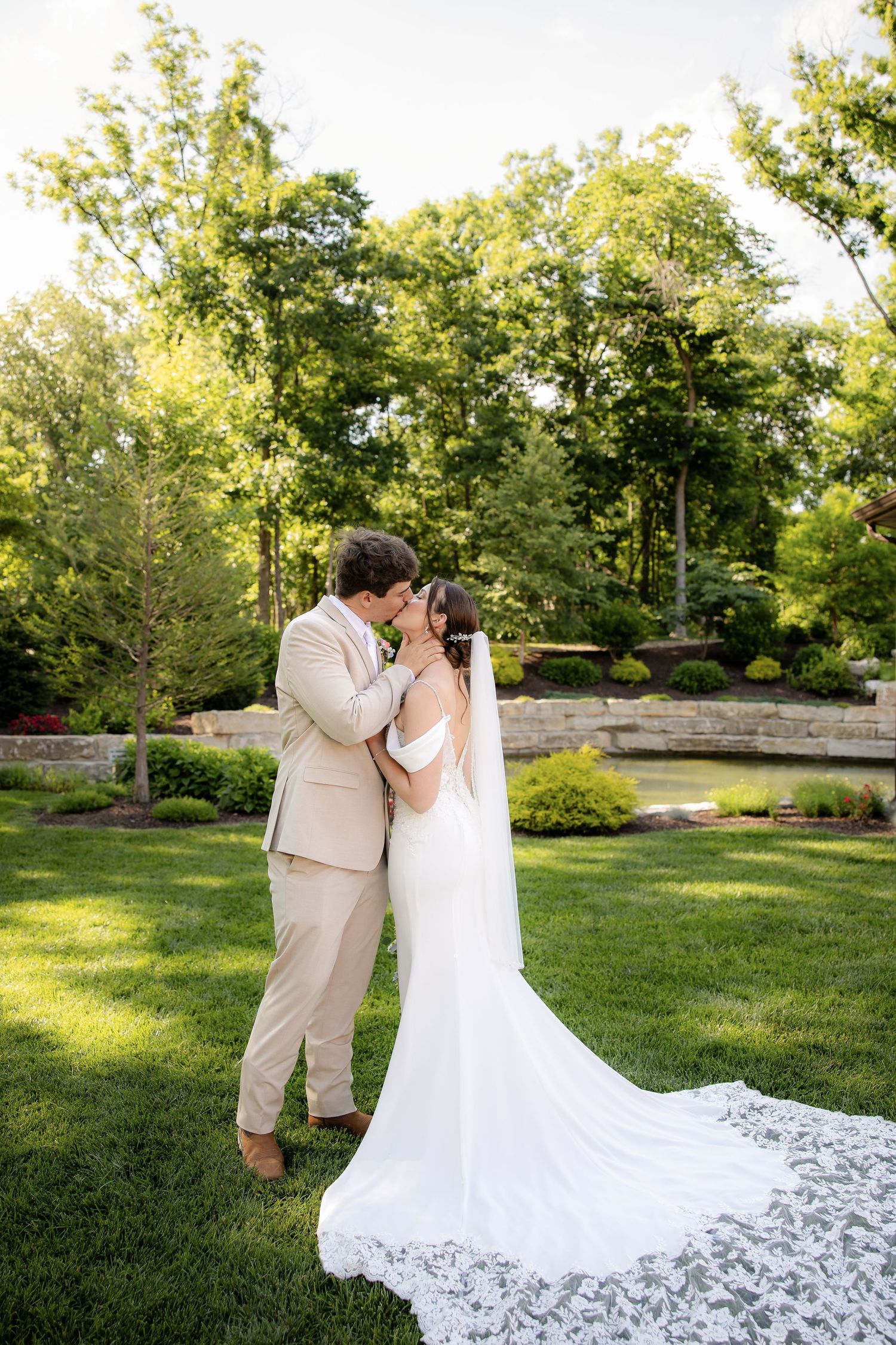 Bride and groom share a romantic kiss in a lush green garden, bride wearing a stunning white gown with lace veil.