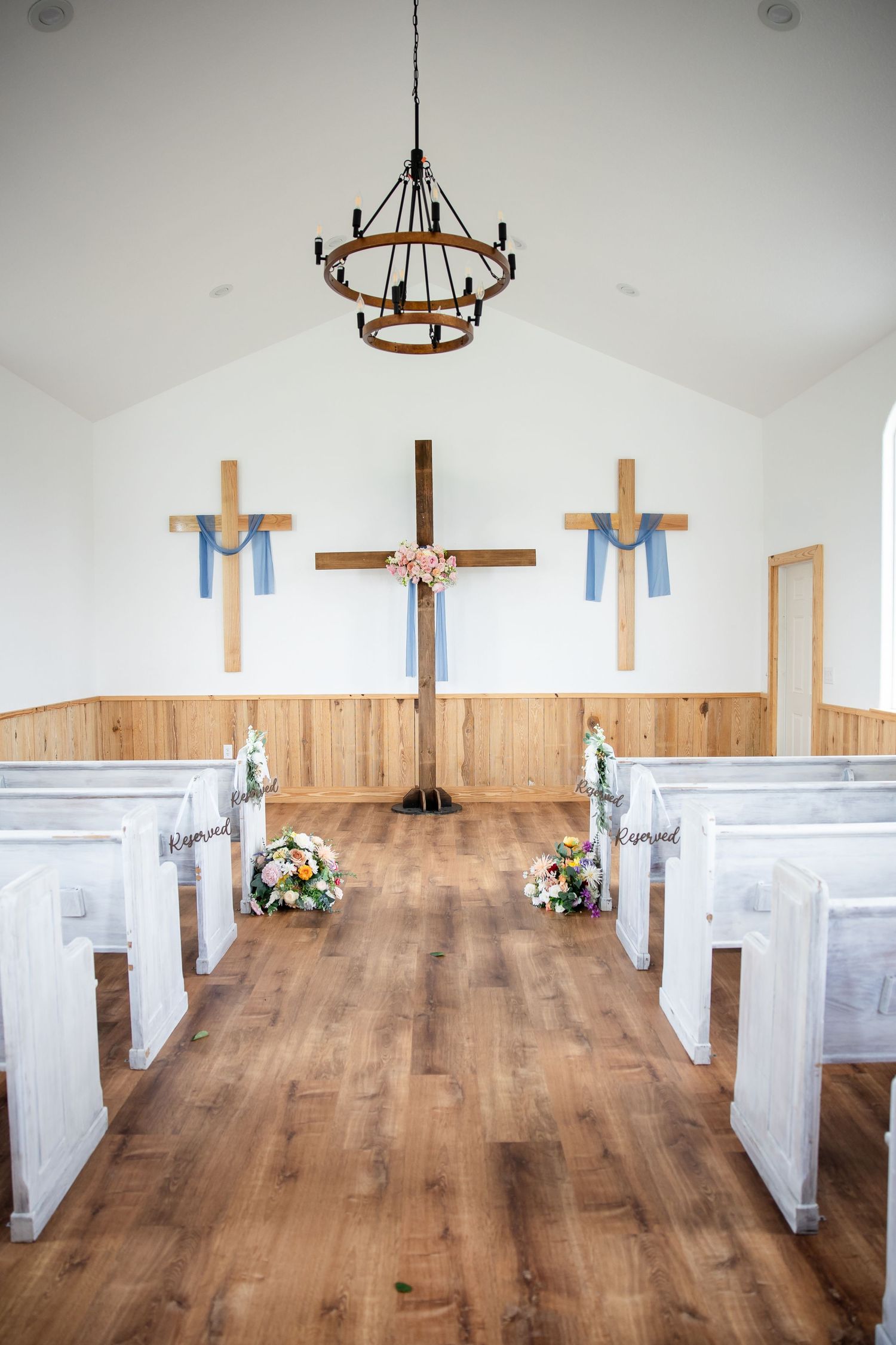 Small chapel interior with wooden crosses, white pews, floral arrangements, hardwood floors, and a rustic chandelier for weddings.