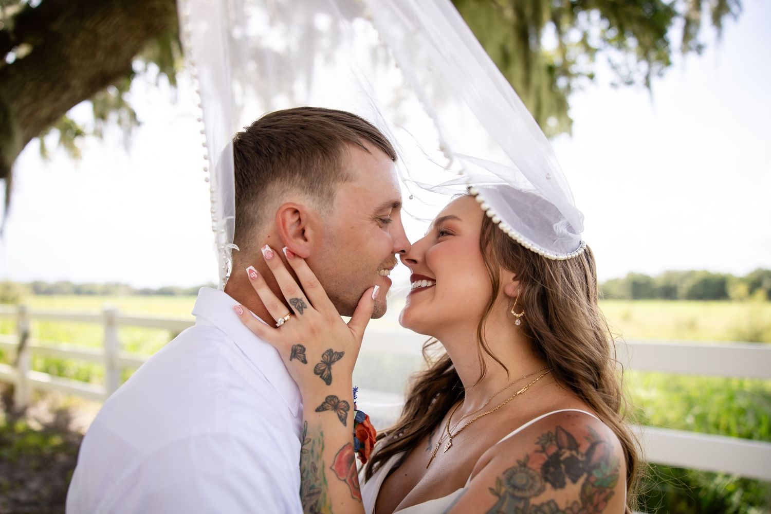 Tattooed bride and groom share a romantic moment under a flowy veil at an outdoor farm wedding.