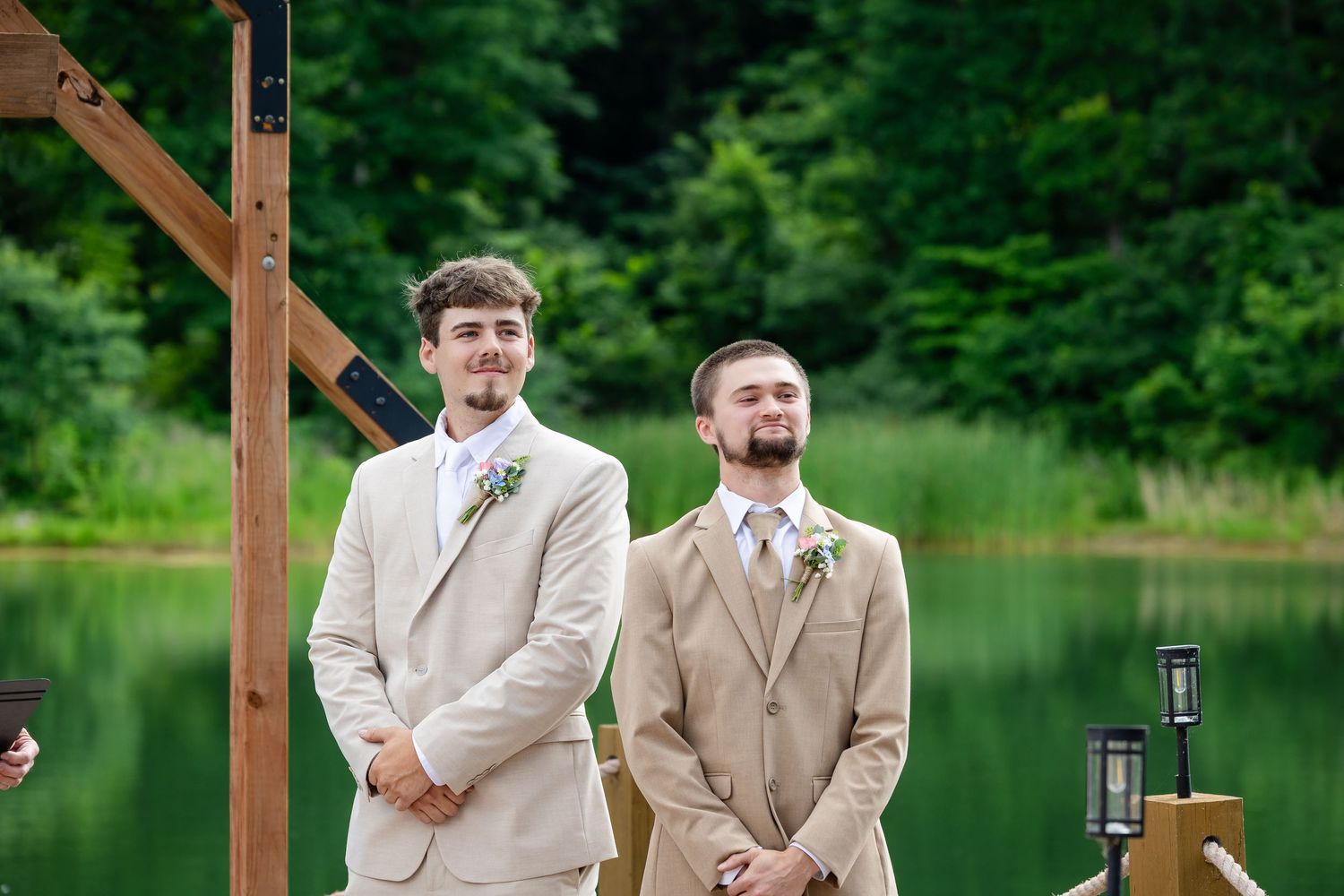 Two groomsmen in tan suits stand at outdoor wedding altar with lush green trees and lake backdrop.