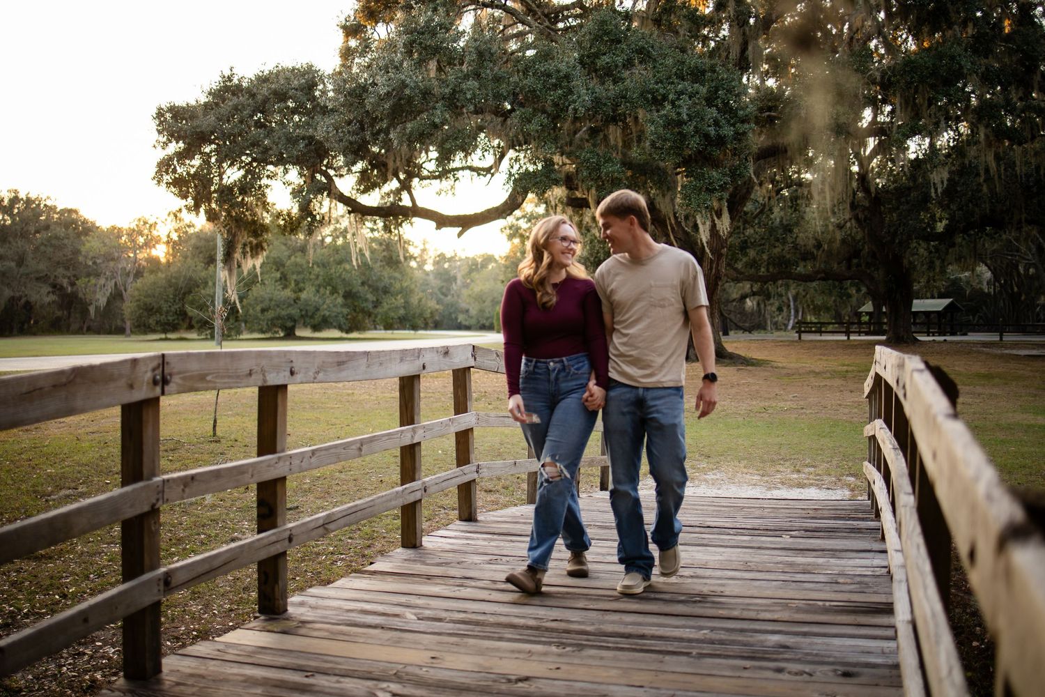 A couple takes a romantic stroll together on a wooden bridge surrounded by oak trees.