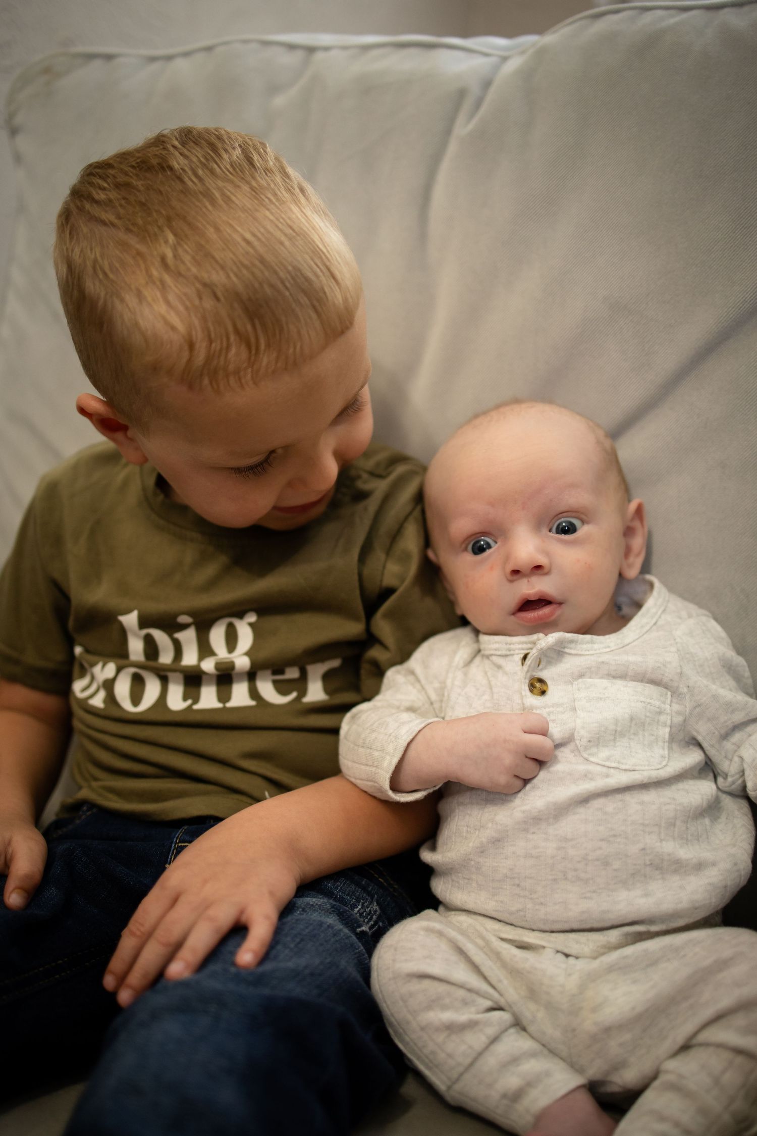 Two children sit together on a light gray couch, with one wearing a brown shirt that says 'big brother'.