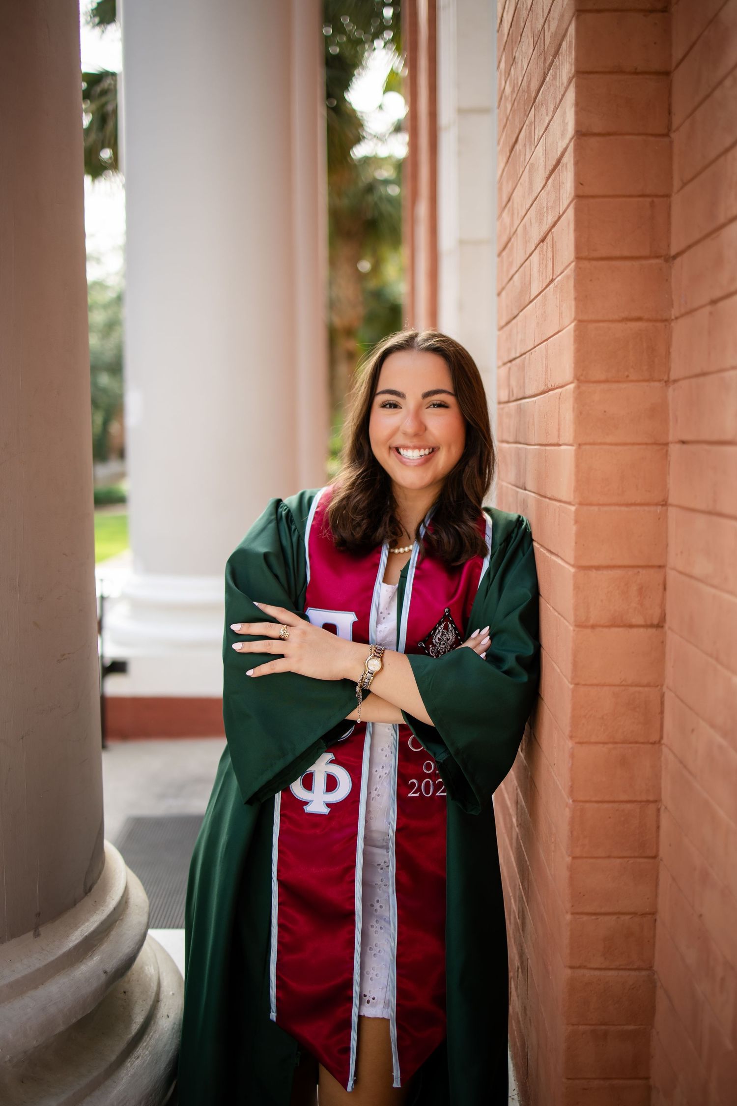 A graduate in a dark green graduation gown and red stole poses while smiling against a brick column outdoors.