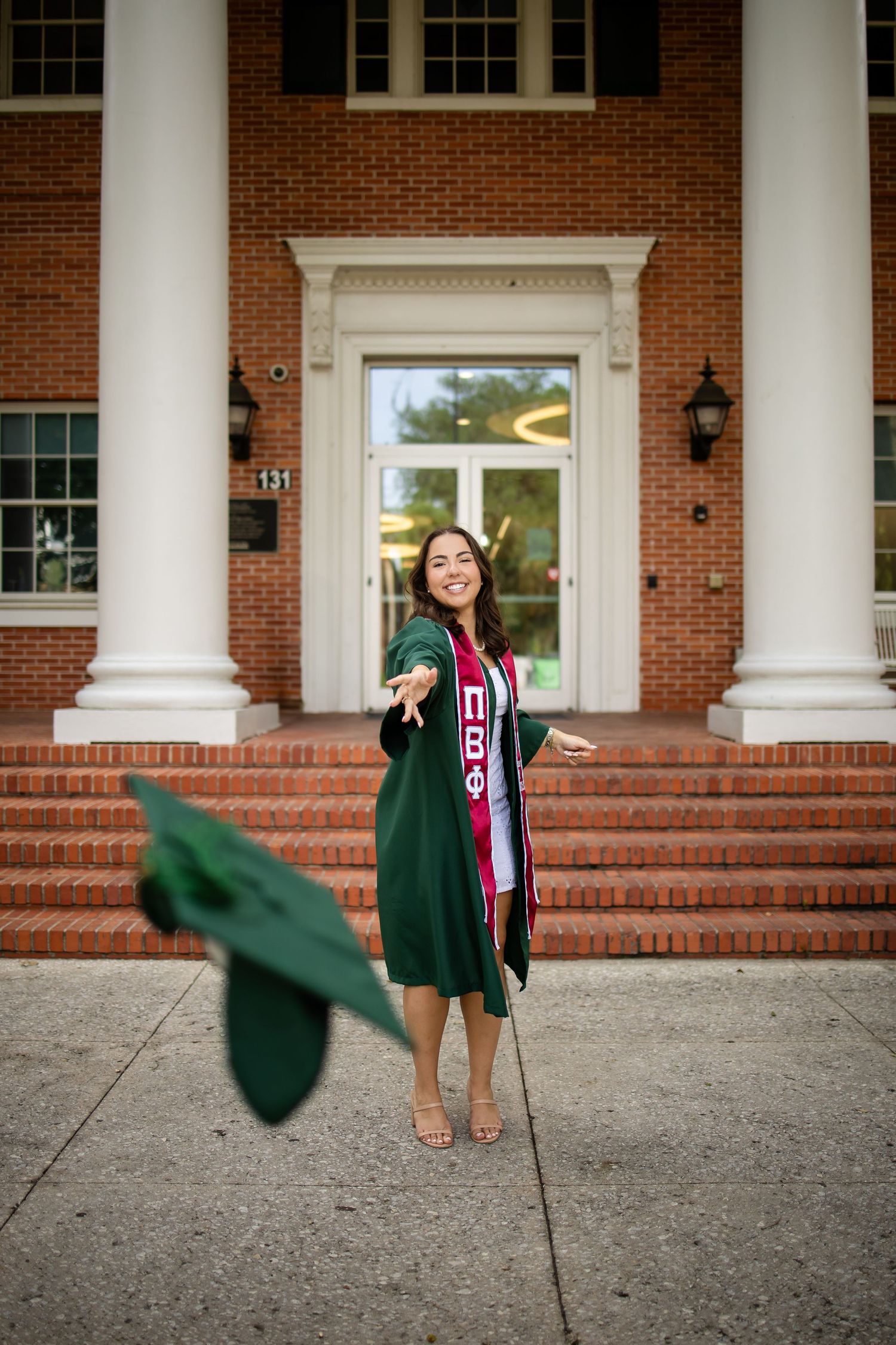 Graduate celebrates by tossing cap while posing in front of collegiate building with white columns.