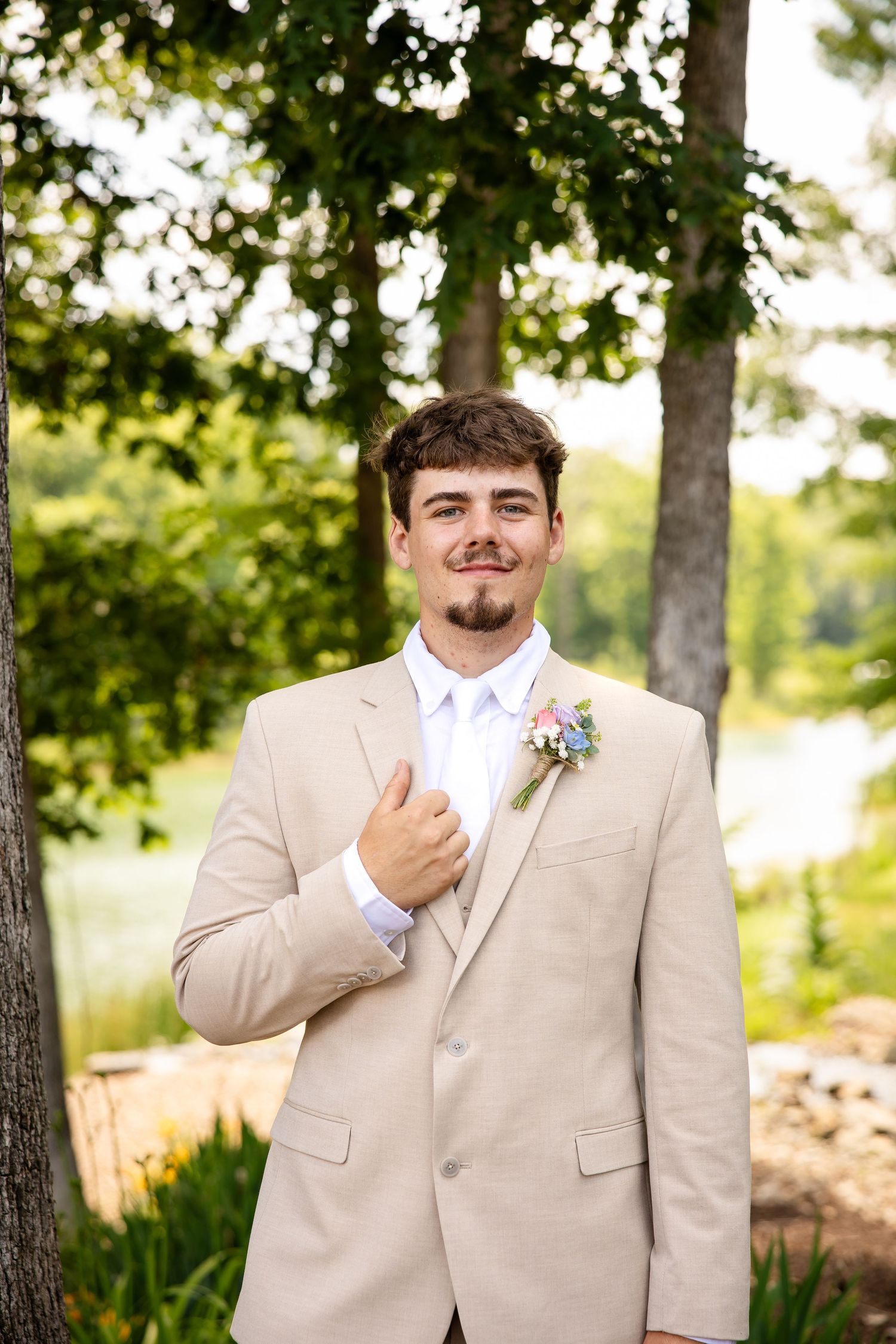 Smiling groom in tan suit with floral boutonniere poses outdoors near trees and a lake on his wedding day.