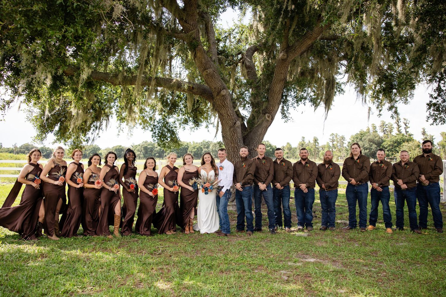 Wedding party poses under a large oak tree, bridesmaids in brown gowns and groomsmen in brown shirts with jeans.