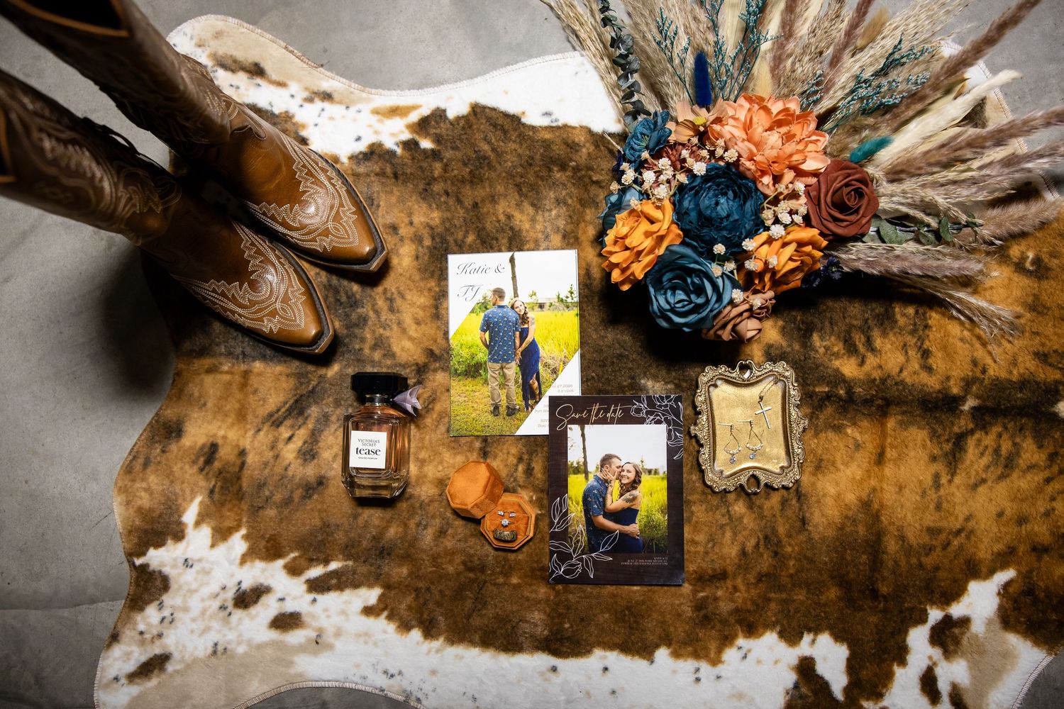 Rustic wedding flat lay with cowboy boots, floral bouquet, photos, and perfume on cowhide rug.