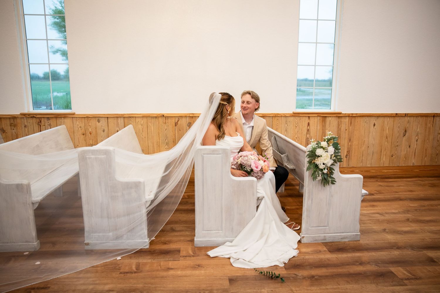Bride and groom sit together in white church pews, bride wearing veil and holding pink bouquet on hardwood floors.