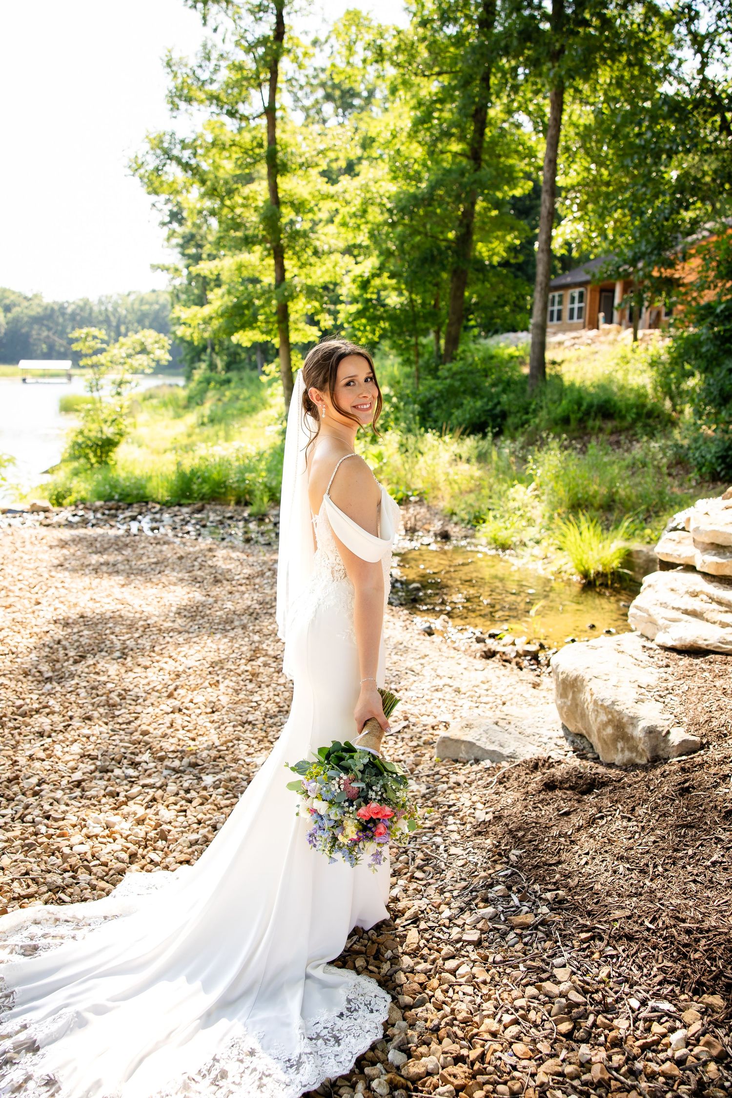 Radiant bride in off-shoulder white gown with lace train poses by a serene lakeside, holding a colorful wildflower bouquet.