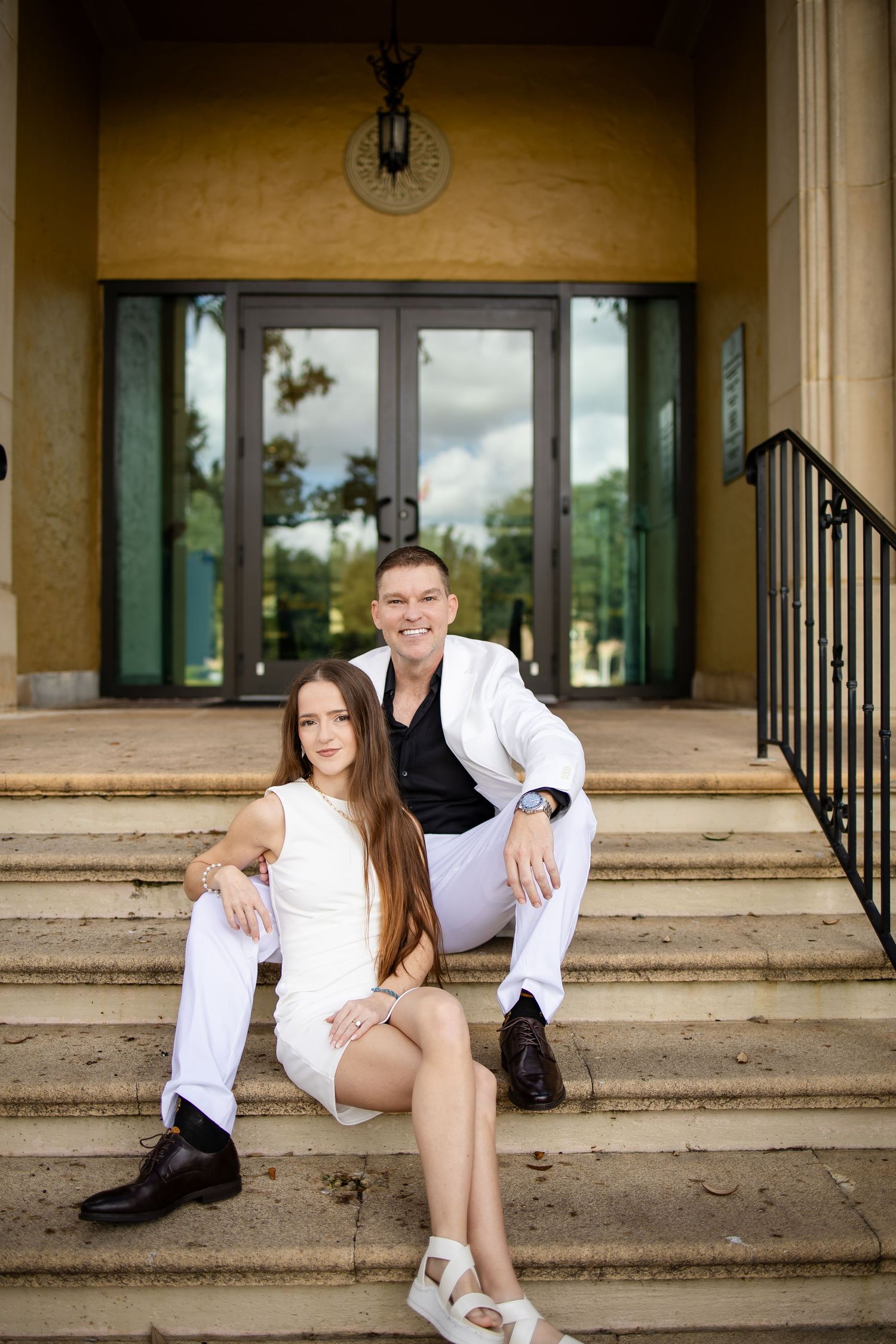 Couple in white outfits sitting on steps of elegant building, smiling for a portrait photo outdoors.