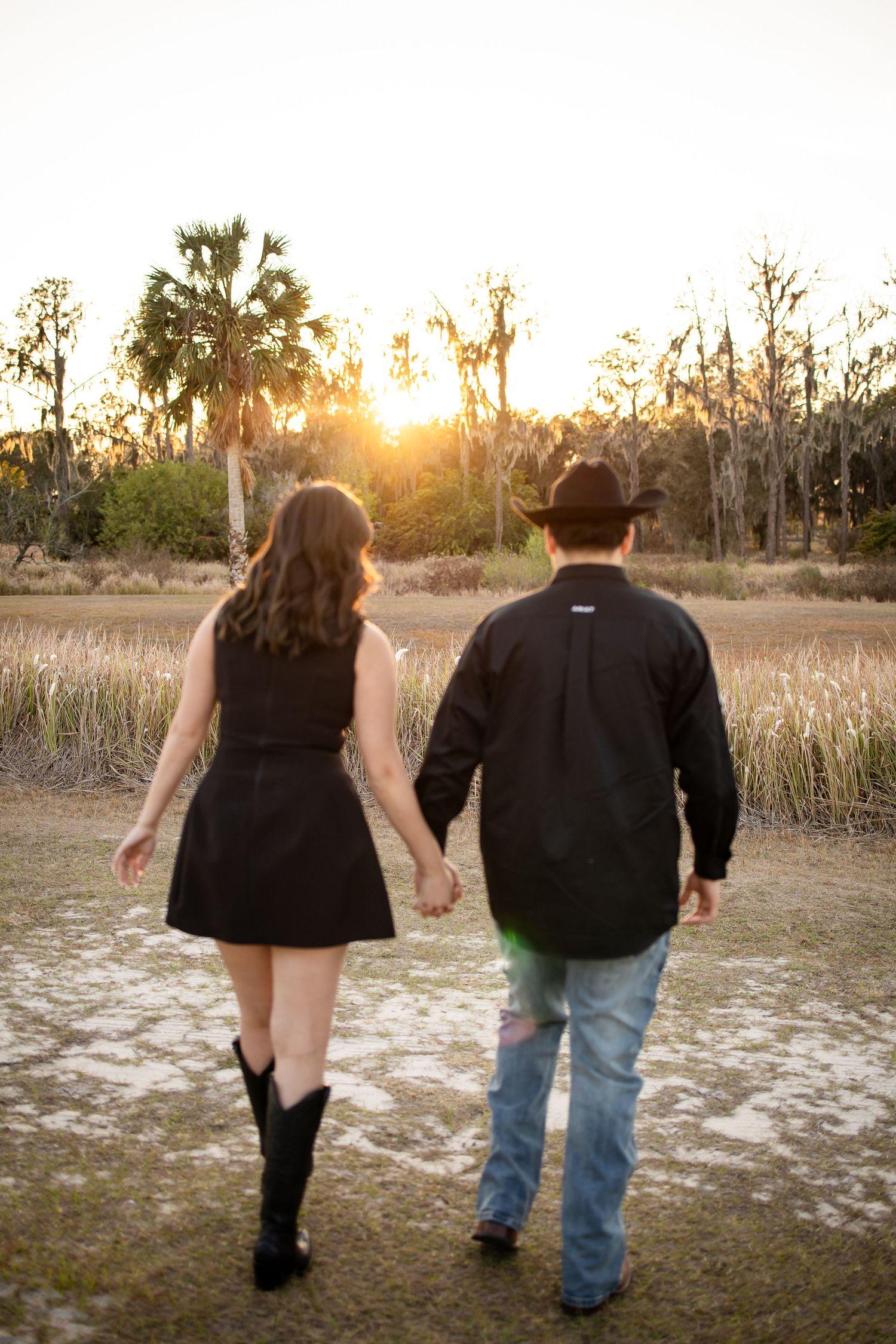 Two people walking hand in hand through a field at sunset with palm trees in the background.