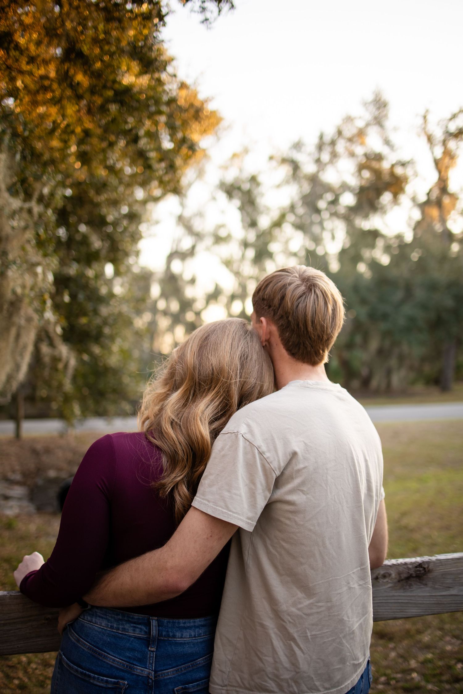 A couple sits close together on a wooden fence at sunset with trees in the background.