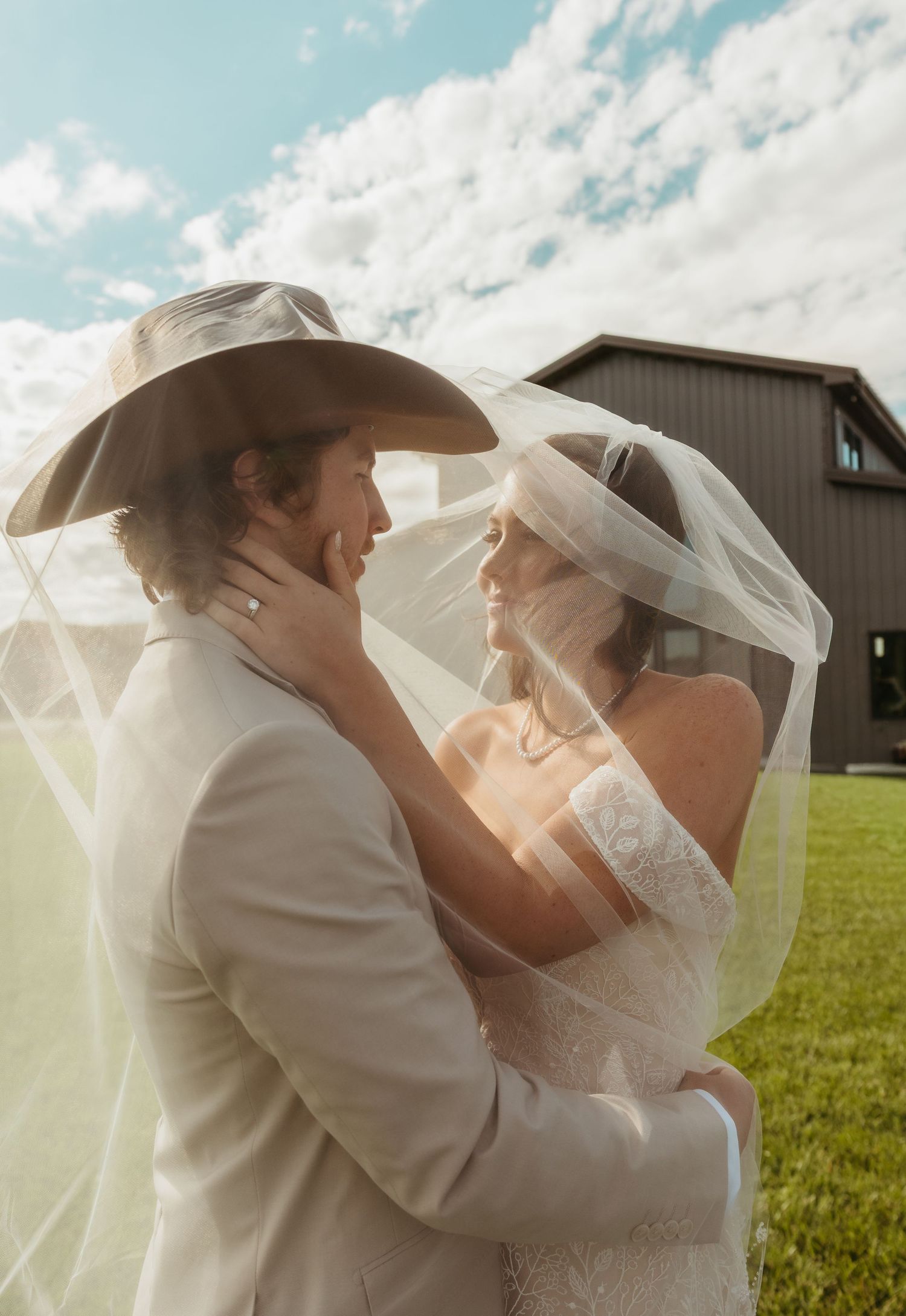 A bride and groom share a romantic moment under a flowing veil outdoors, groom wearing a cowboy hat, barn in background.