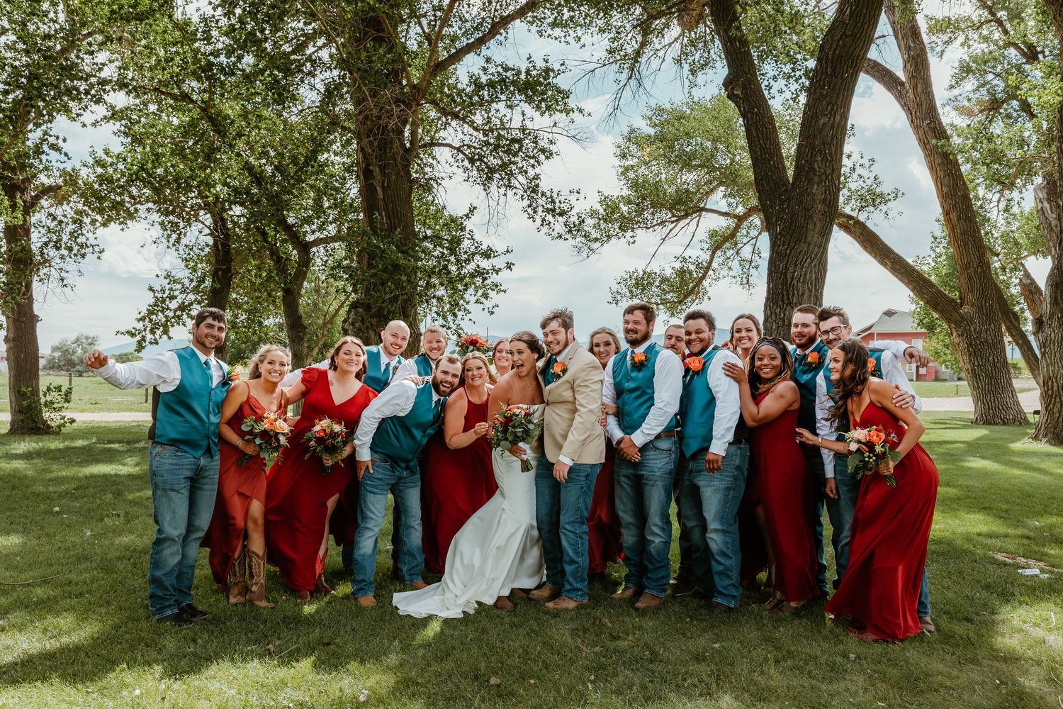 Wedding party in red dresses and teal vests poses outdoors under large trees on a sunny day.
