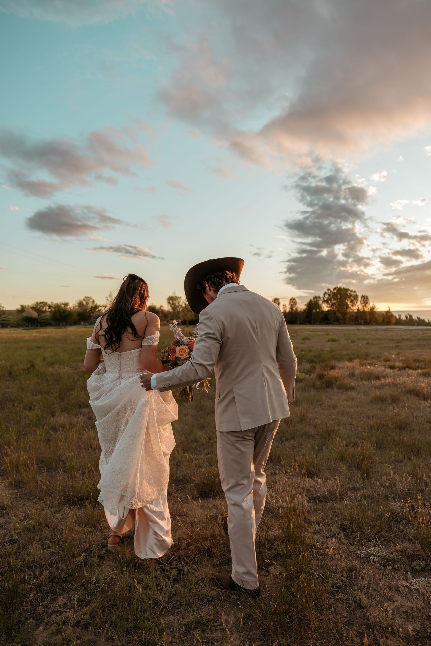 Bride and groom walking through a golden field at sunset, bride in white gown, groom in tan suit and cowboy hat.
