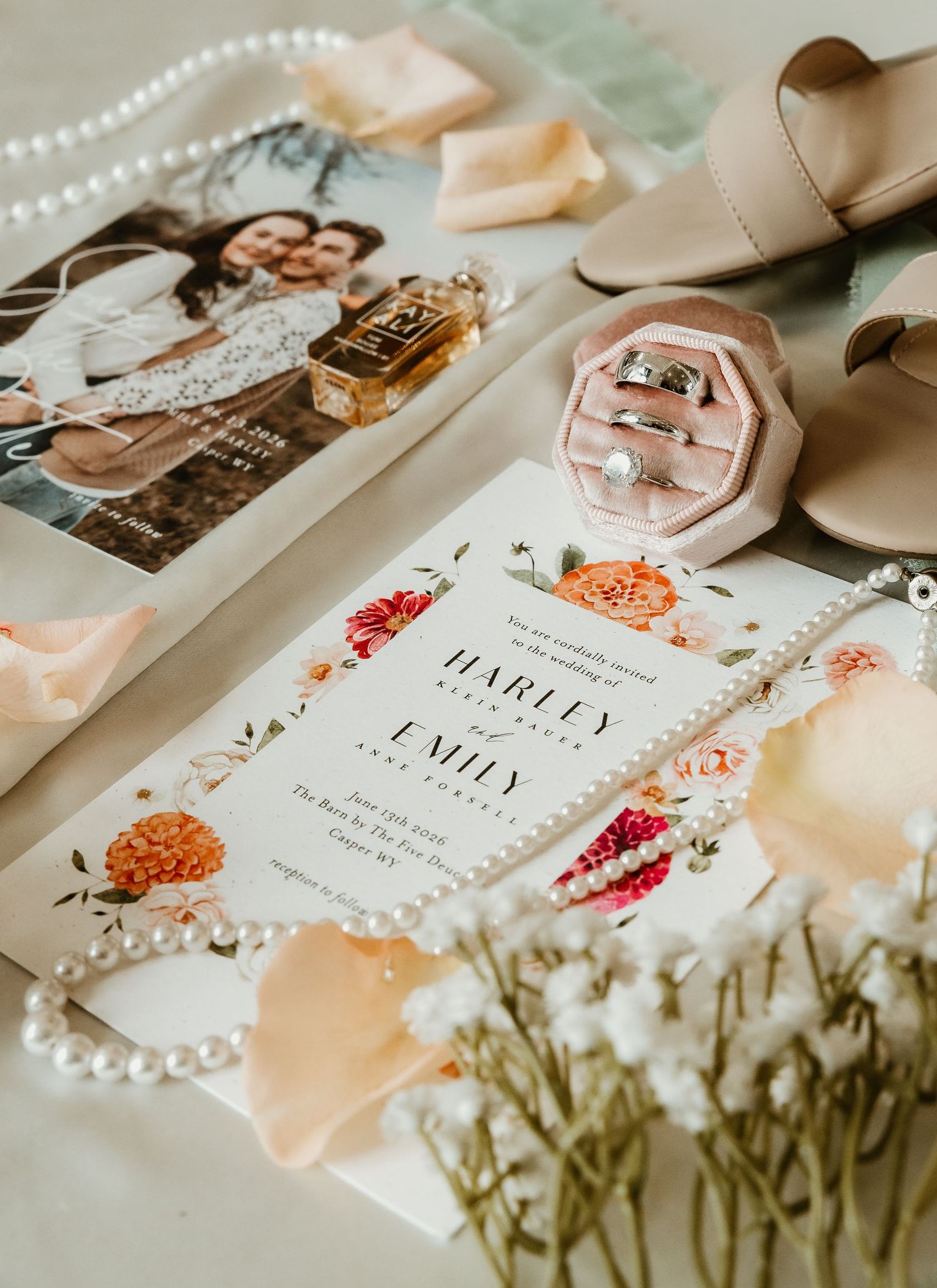 Elegant wedding flat lay with floral invitation, pearl necklace, rings in pink velvet box, perfume, photo, and bridal heels.