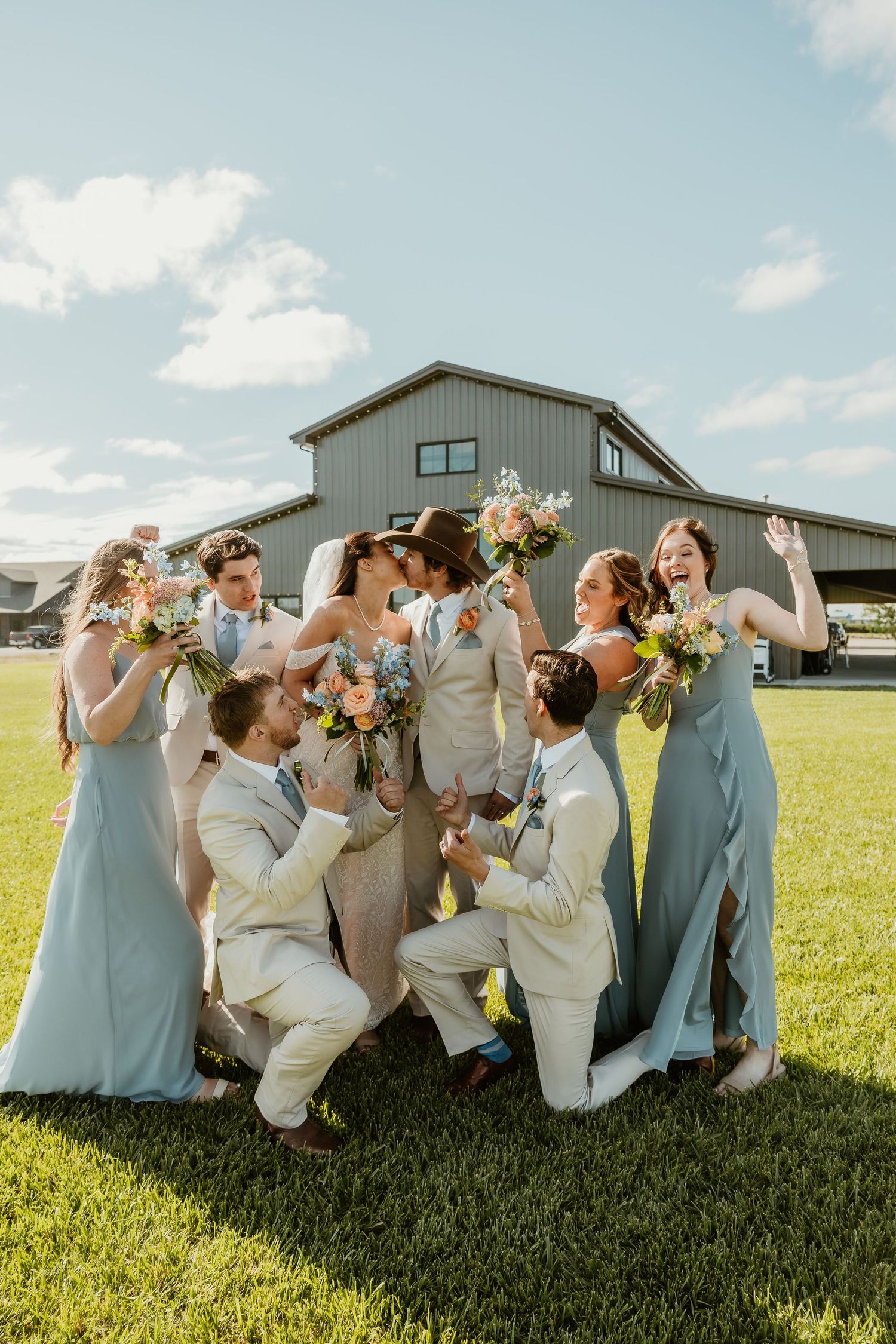 Joyful wedding party in dusty blue gowns and cream suits celebrating outdoors near a barn on a sunny day.