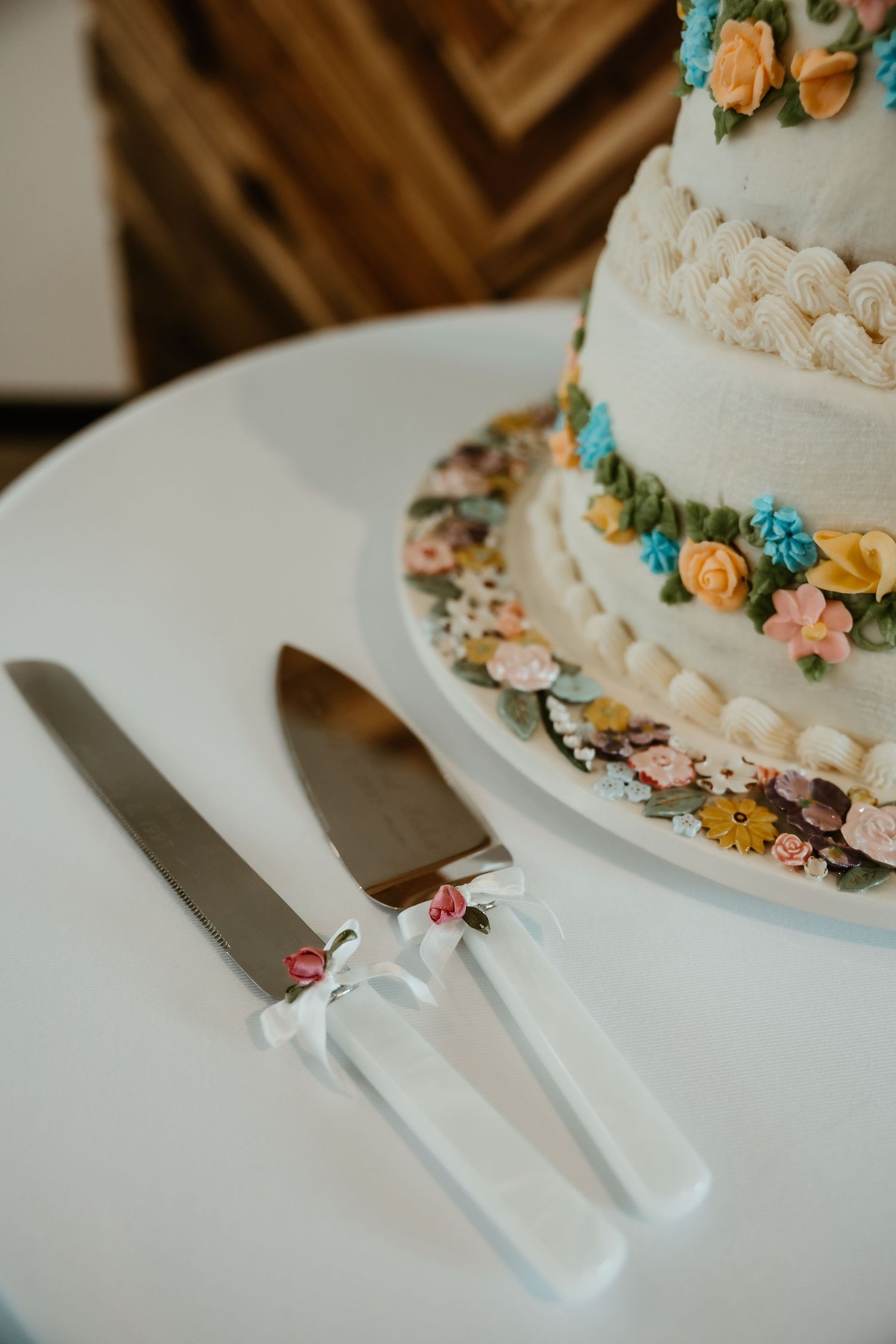 White wedding cake with colorful floral decorations beside elegant cake knife and server set with white bows on a round table.