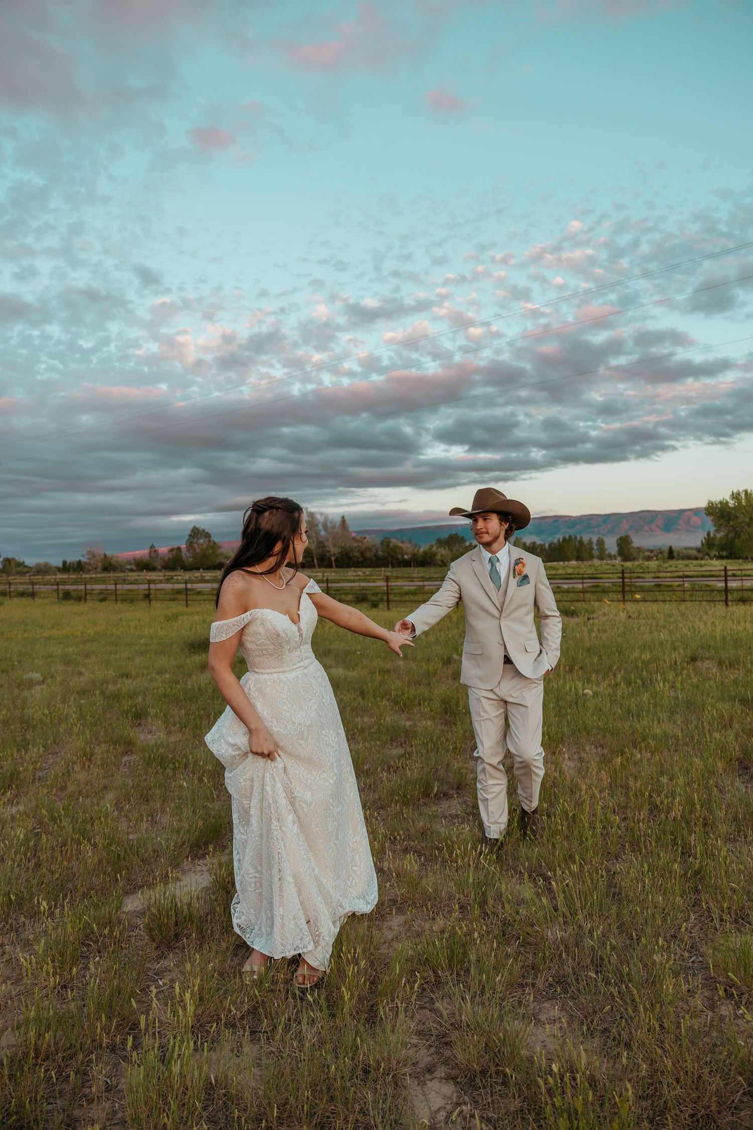 Bride and groom holding hands in a green field at sunset, wearing a white gown and tan suit with cowboy hat.