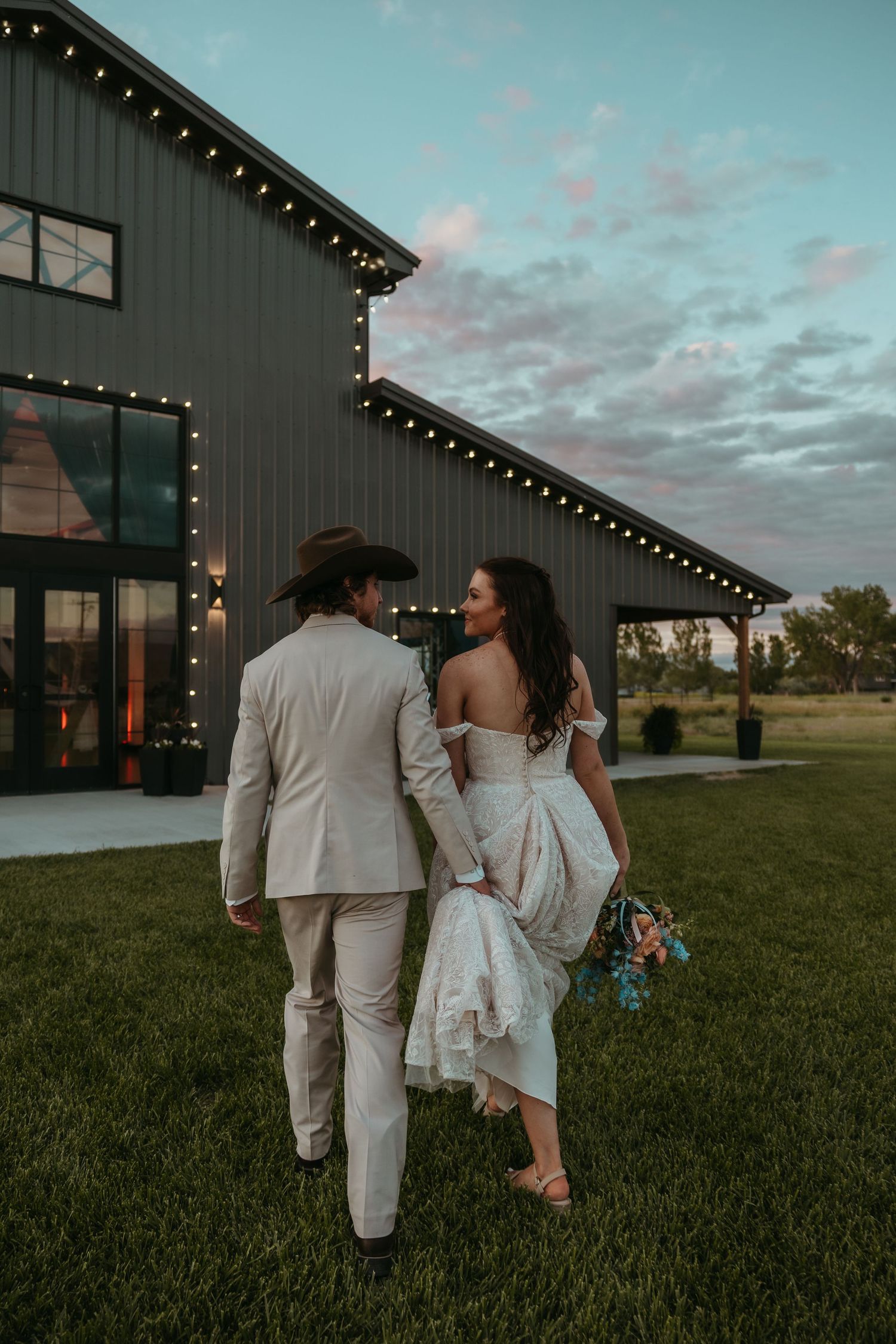 A bride and groom walk hand-in-hand toward a barn venue at dusk, string lights glowing against a pastel sunset sky.
