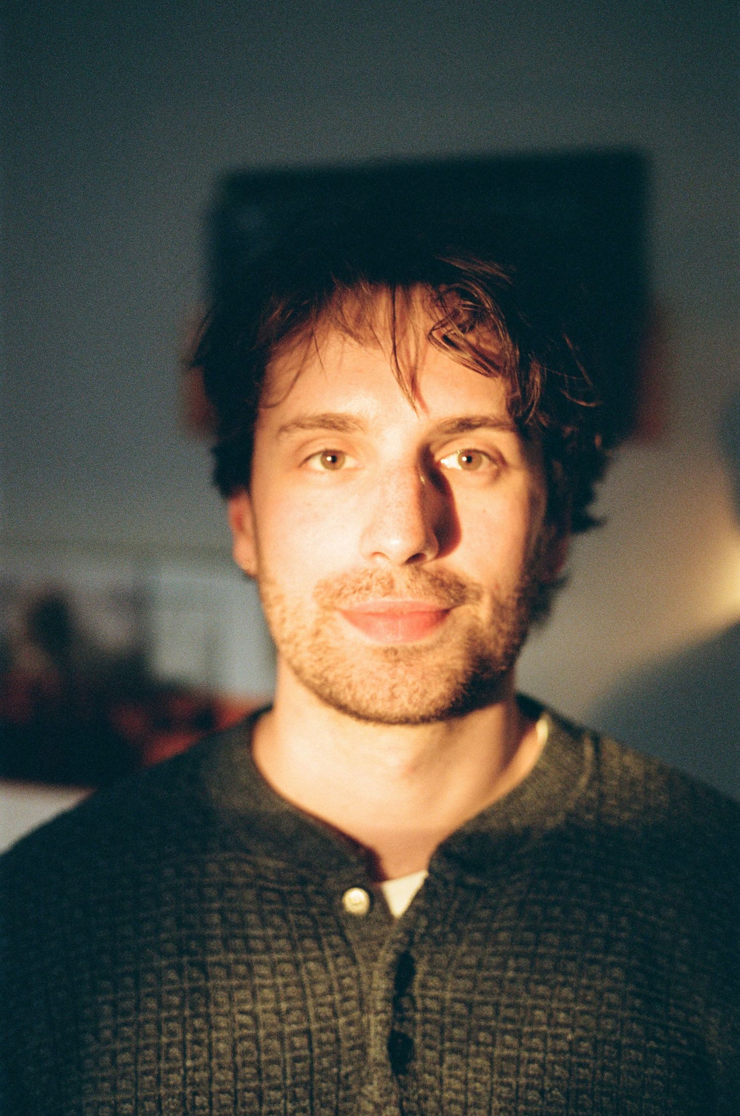 Young man with curly brown hair and stubble wearing a dark green henley sweater, lit by warm natural light indoors.