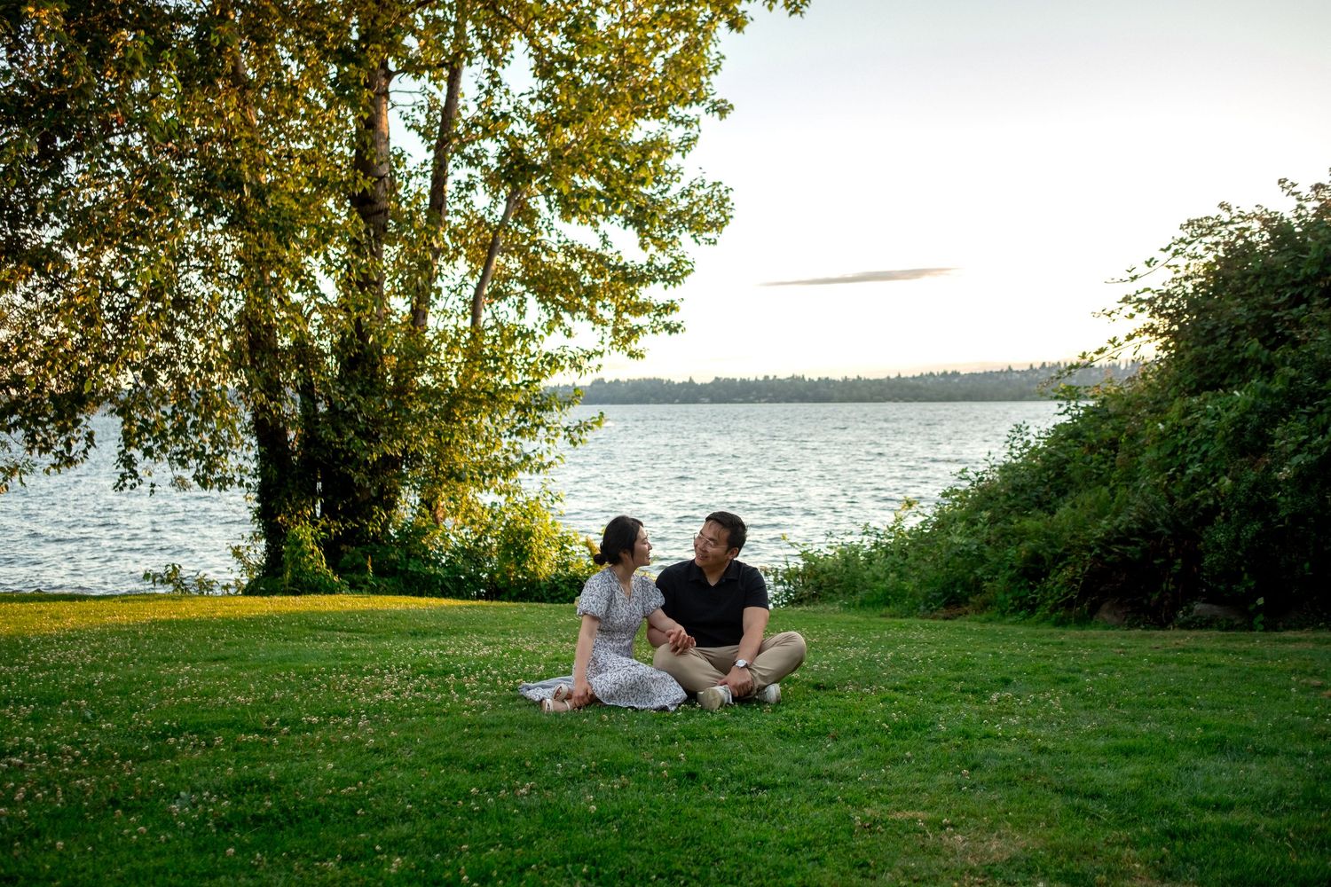 Couple sitting on green grass by a lake at sunset, surrounded by trees in a peaceful outdoor setting.