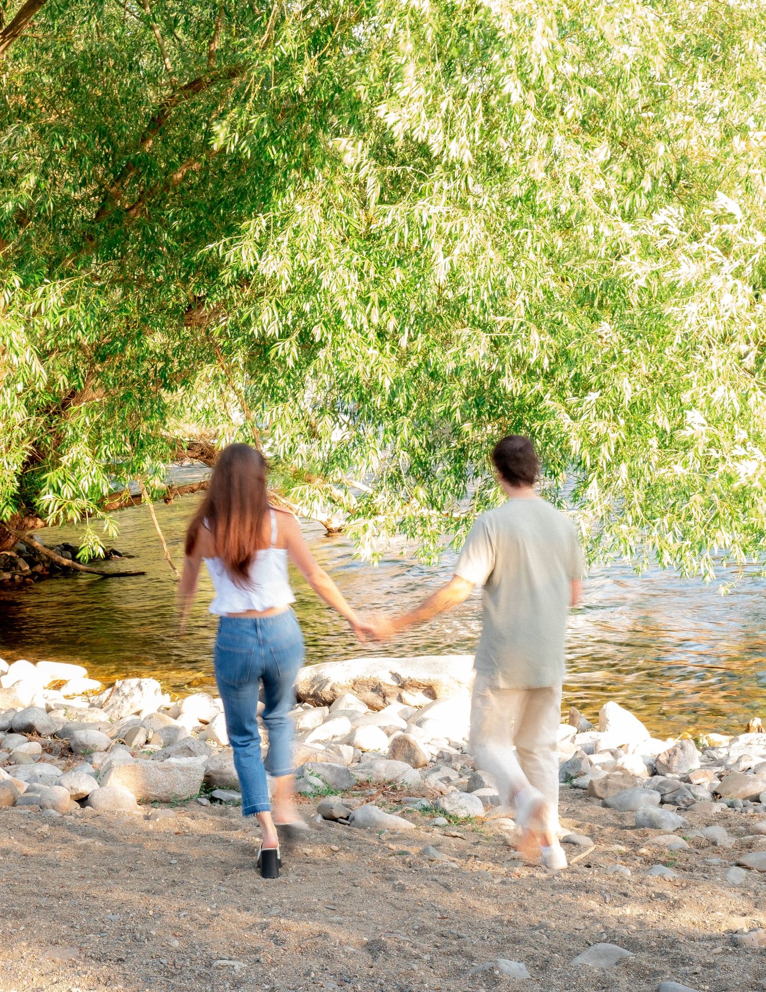 Couple holding hands while walking along rocky riverbank under weeping willow trees.
