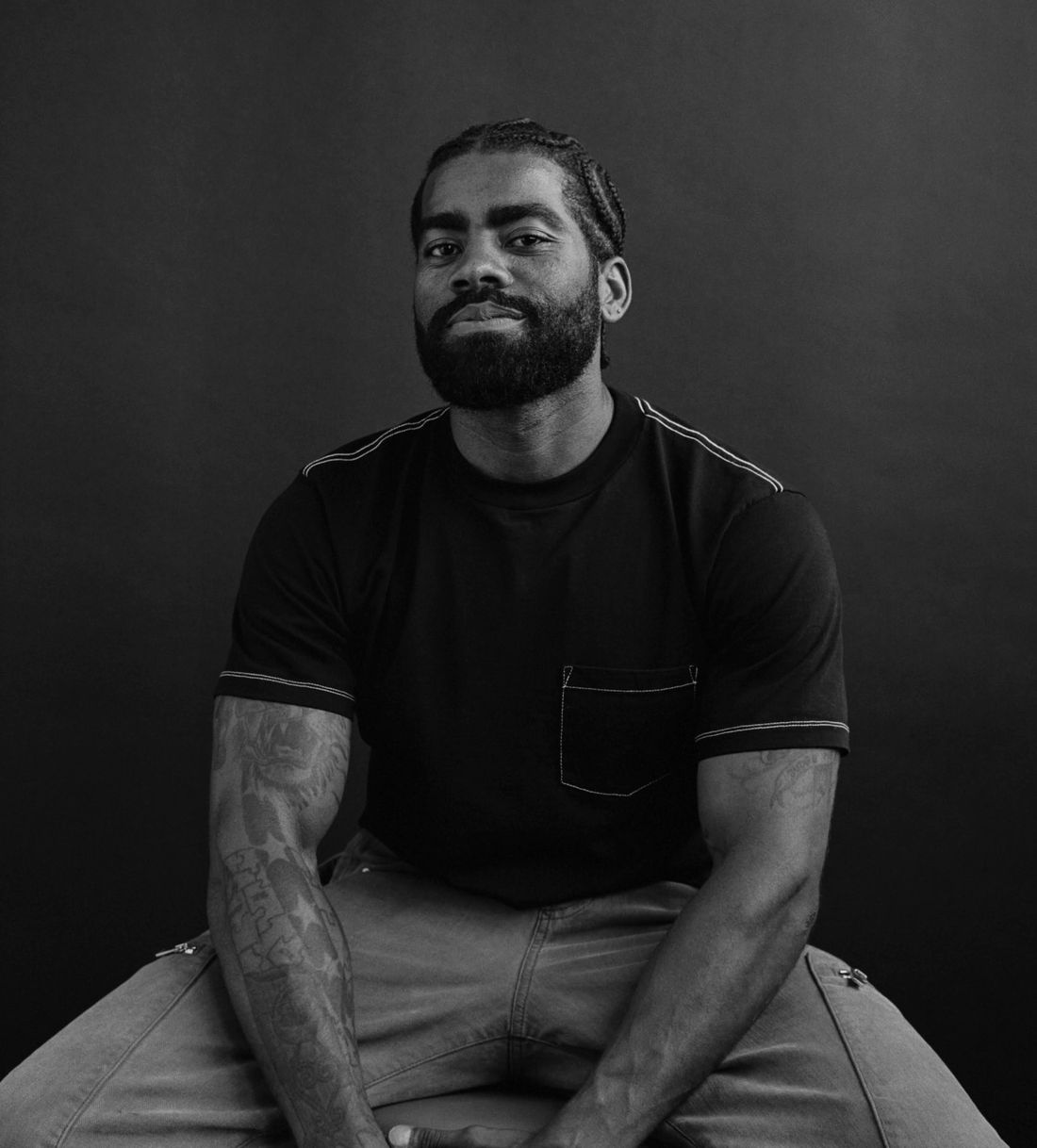 Black and white portrait of a bearded man with tattoos sitting confidently in a black t-shirt against a dark background.