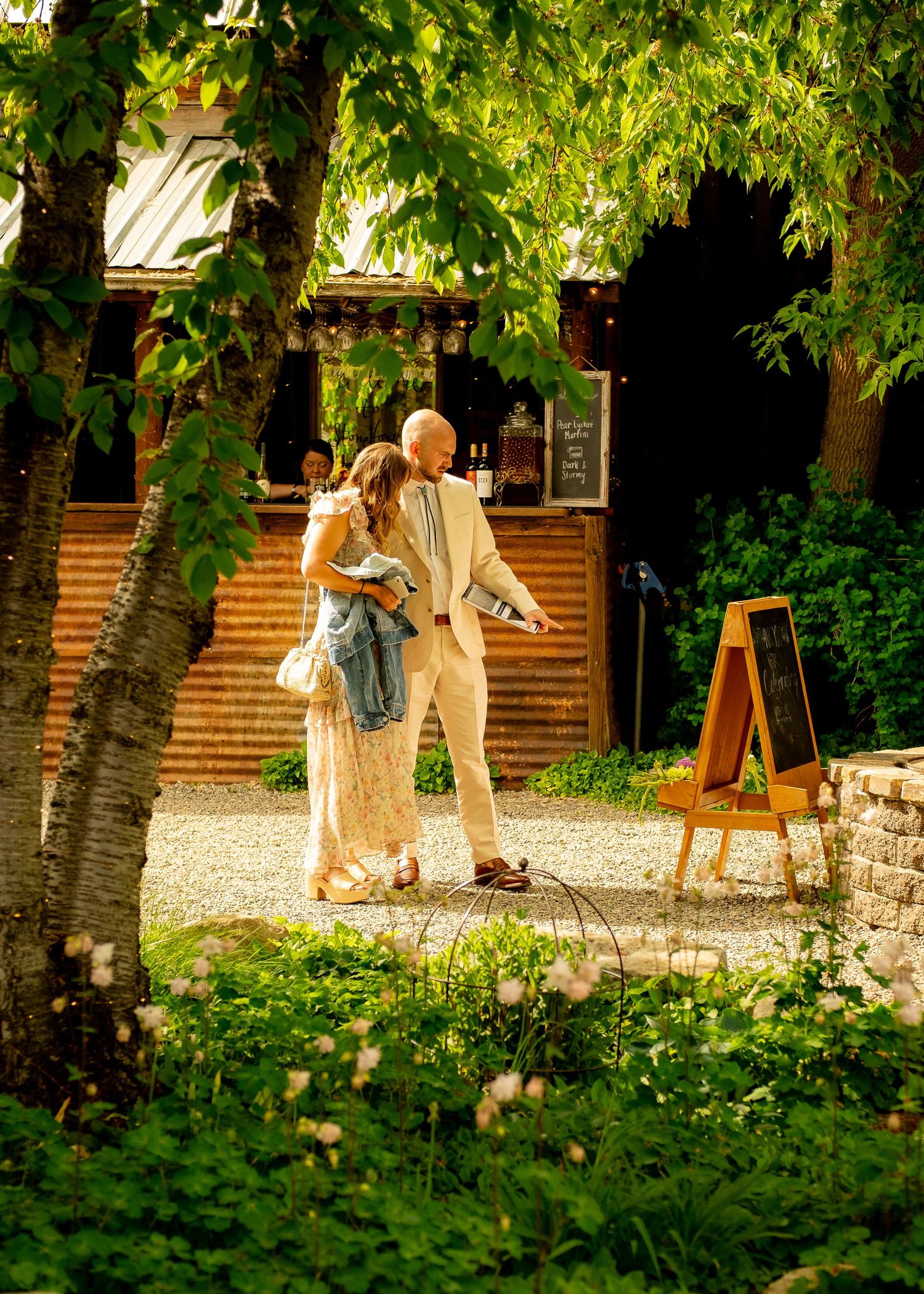 Artists paint outdoors in a lush garden setting with wooden buildings and gravel paths on a sunny summer day.