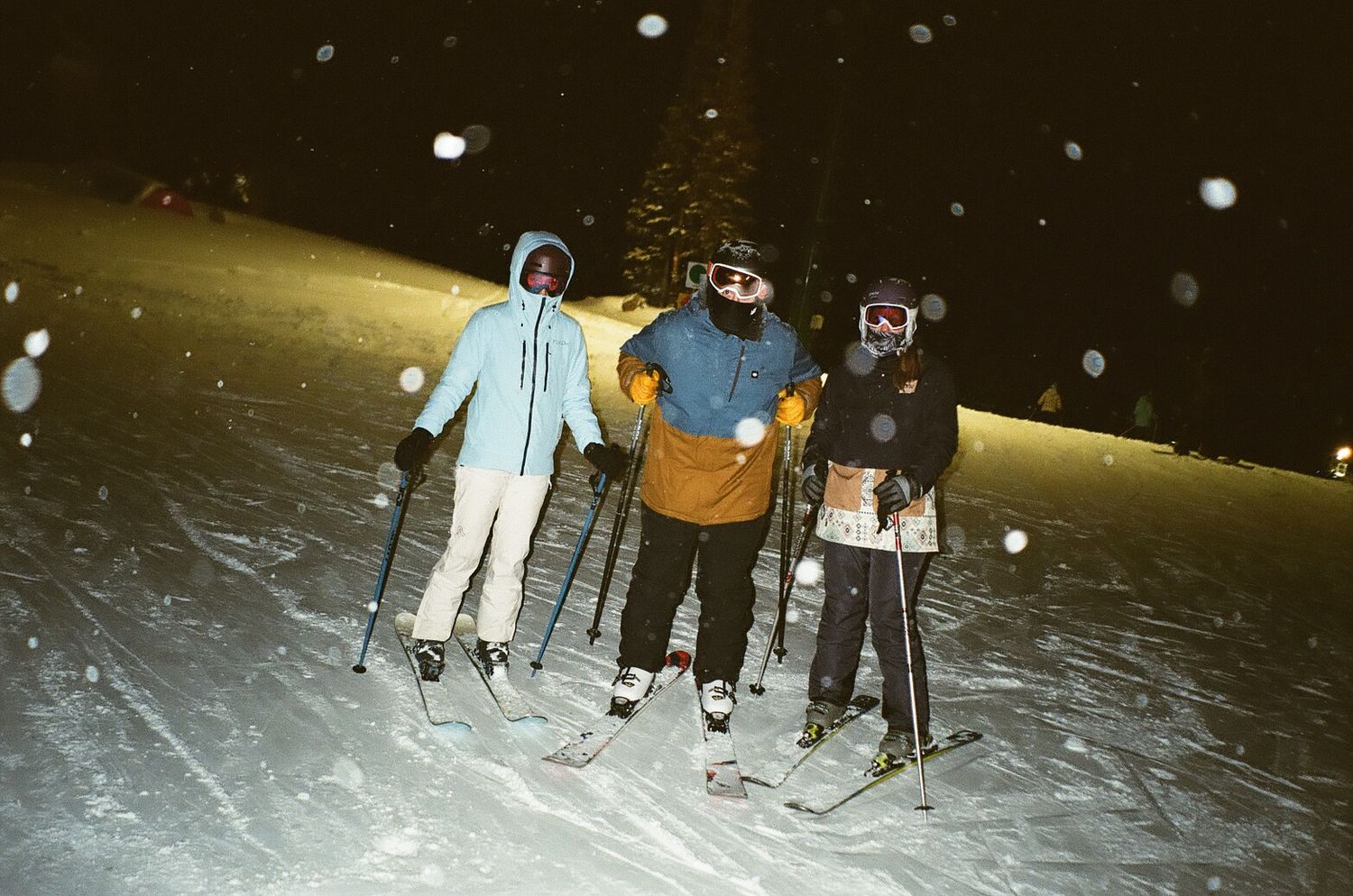 Three people night skiing on a snowy slope with falling snow visible in the light.
