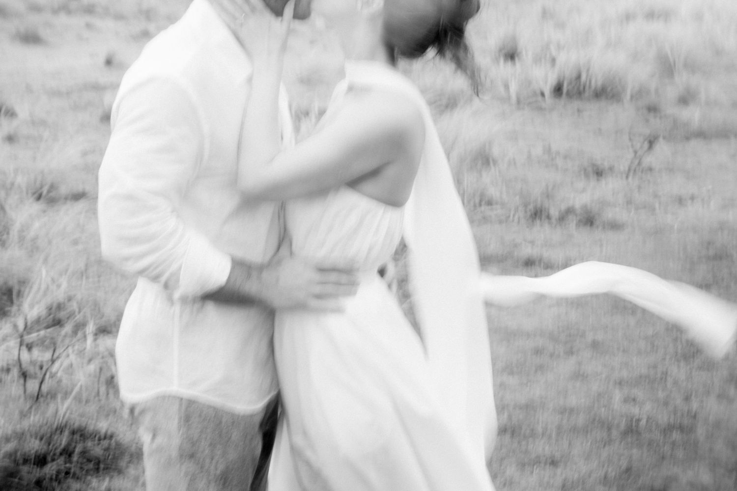 Black and white photo of a couple embracing and kissing outdoors in a field, bride in flowing white dress.
