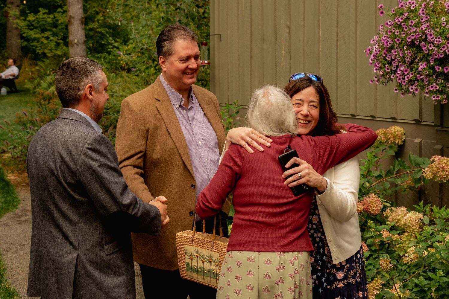 Group of people socializing outdoors, two women hugging warmly near flowering plants and a green barn.