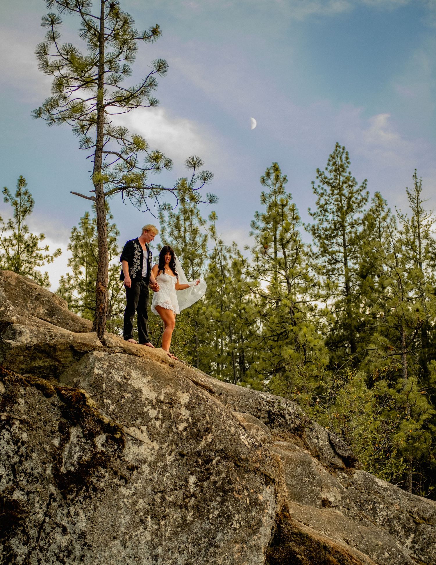Couple standing on a rocky outcrop surrounded by tall pine trees under a blue sky with wispy clouds.