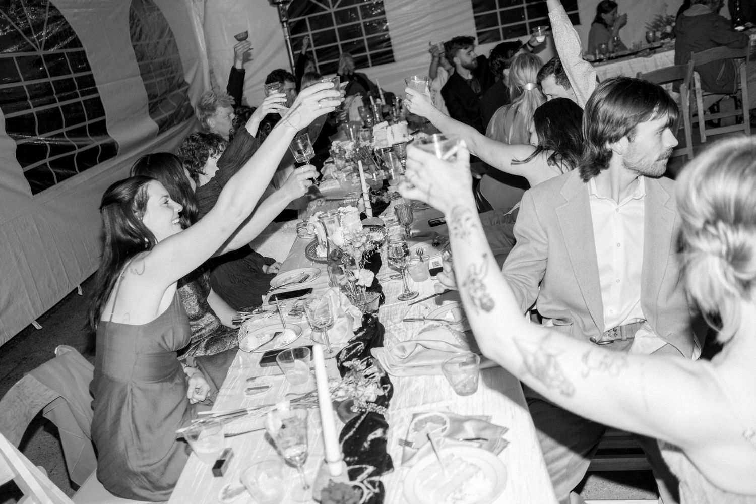 Black and white photo of wedding guests raising glasses in a toast at a long decorated reception dinner table.