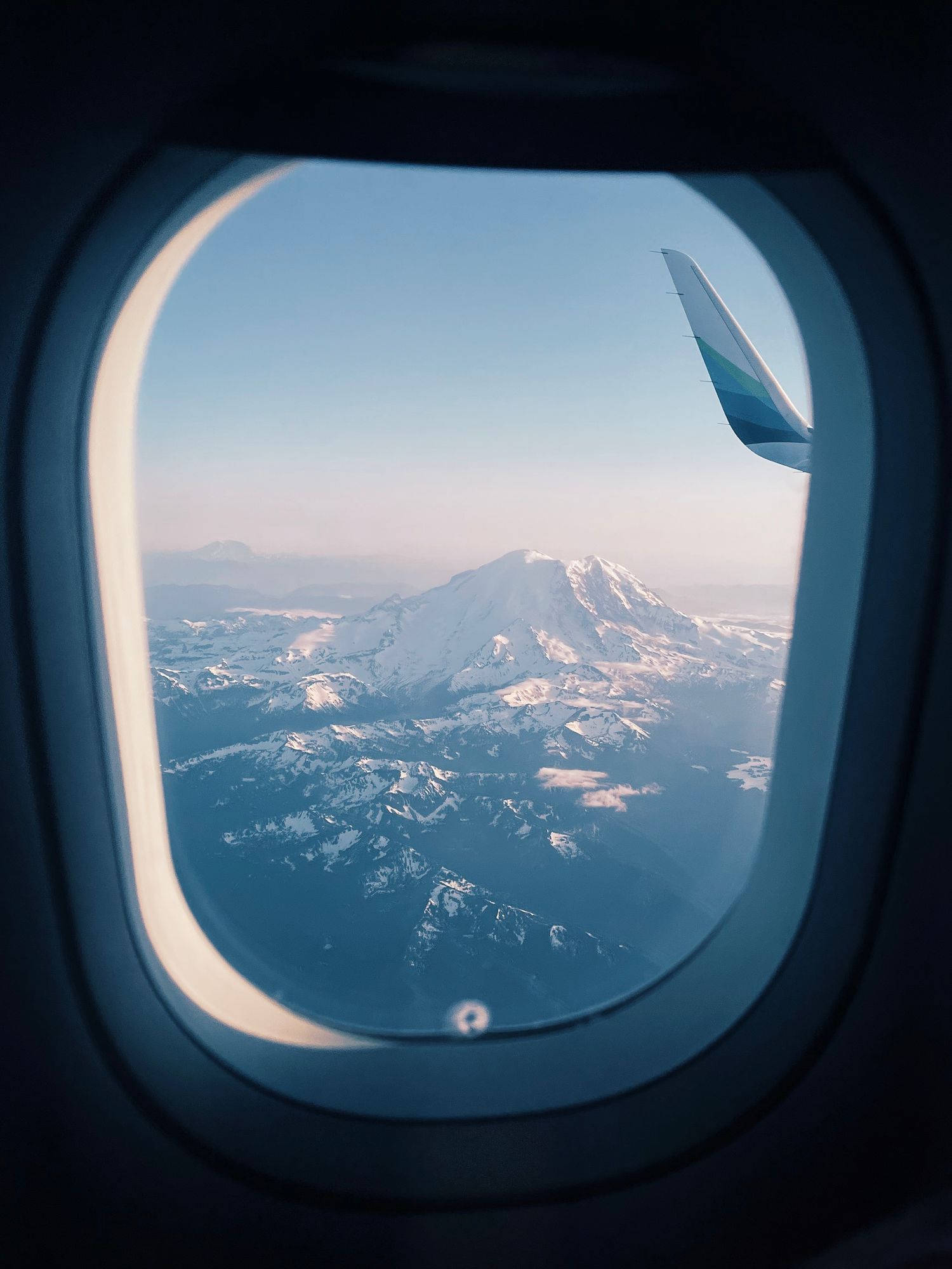 Snow-capped mountain peak viewed through airplane window with wing visible during sunset flight.