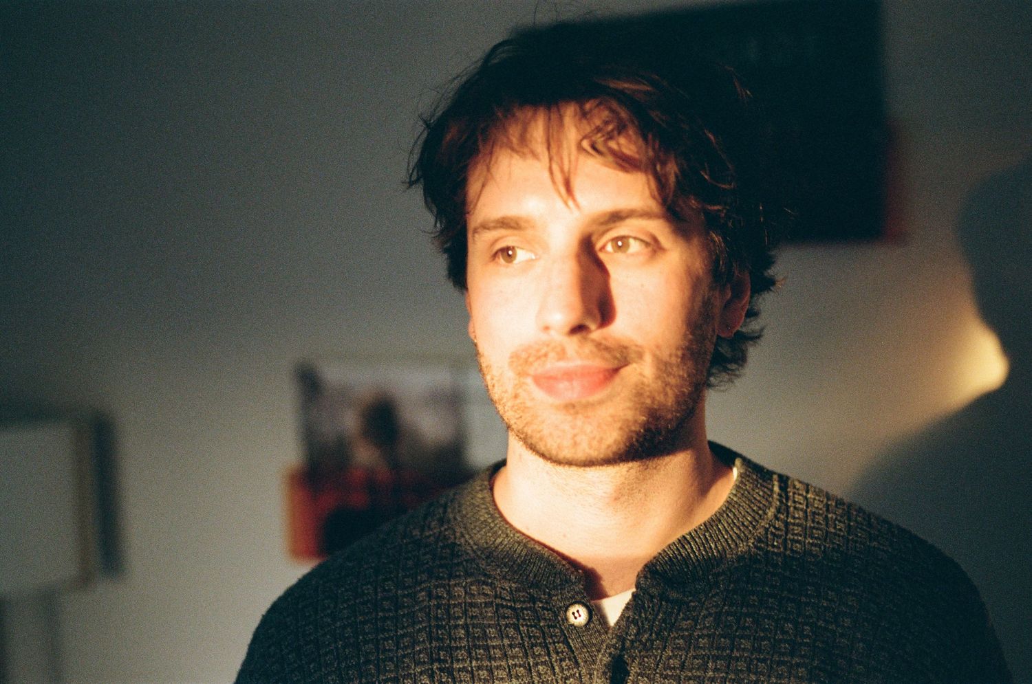 Young man with curly hair wearing a dark sweater, photographed indoors in warm natural light.