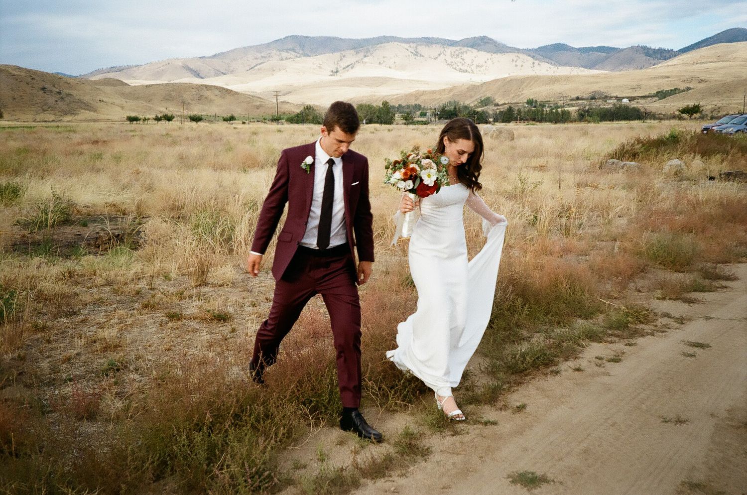 A couple in wedding attire walks along a dirt path through golden hills and mountains in a scenic outdoor setting.