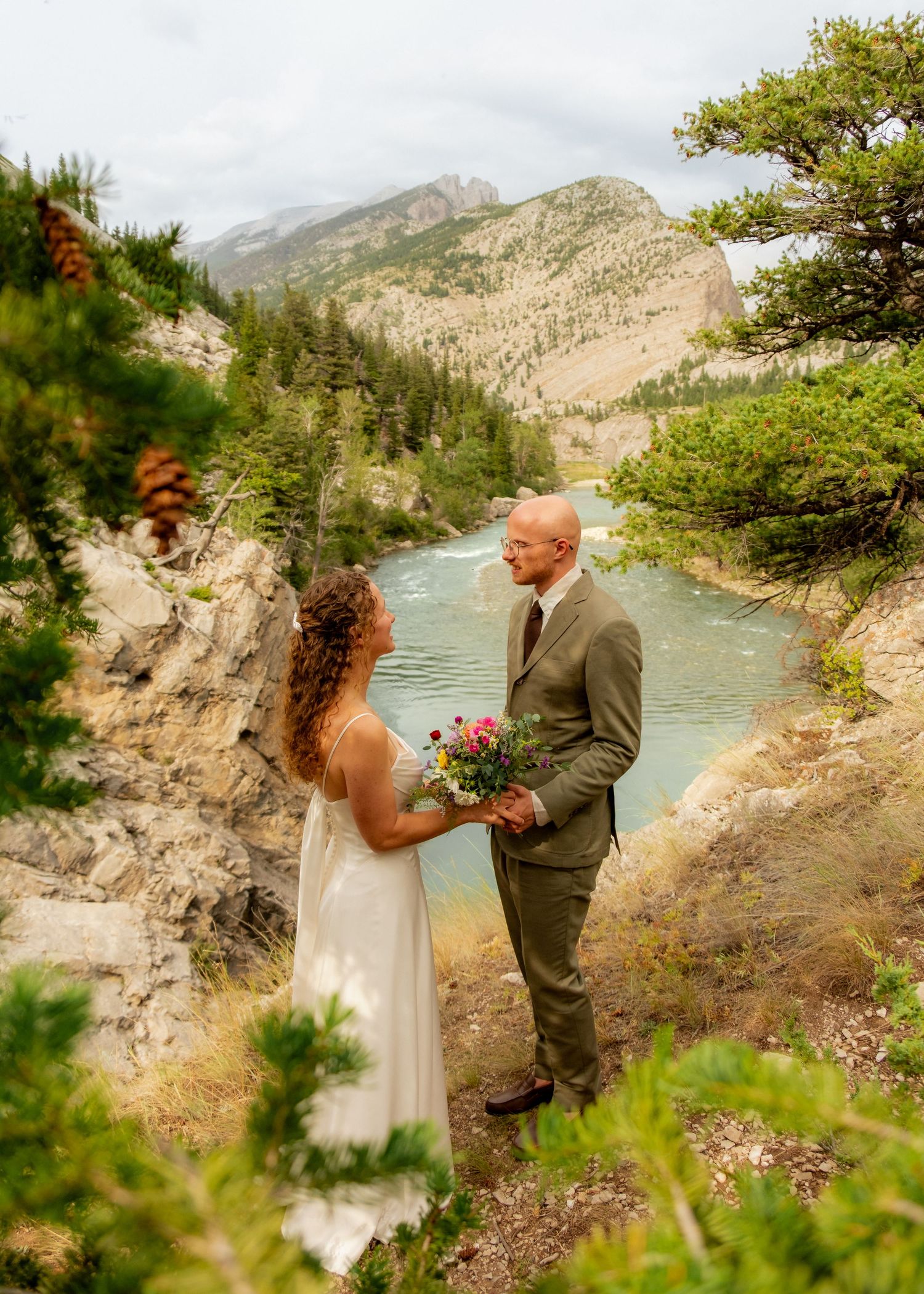 Couple exchanging vows outdoors on rocky cliffs with turquoise river and mountain backdrop.