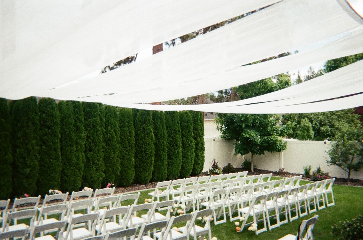 Outdoor wedding ceremony setup with white chairs and fabric canopy in a garden setting.