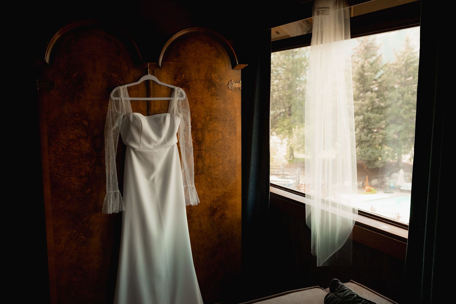 A white wedding dress hangs elegantly by a window in a rustic wooden room with natural light.